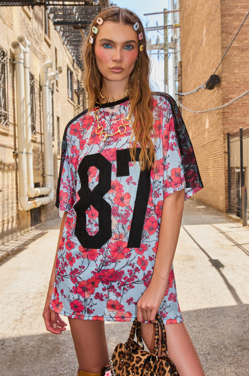 A woman stands in a sunlit alley wearing the POPPIN POPPIES LACE JERSEY DRESS, an oversized sporty style with a bold black “87” over a pink floral print. She accessorizes with colorful hair clips and carries a leopard print bag. 0