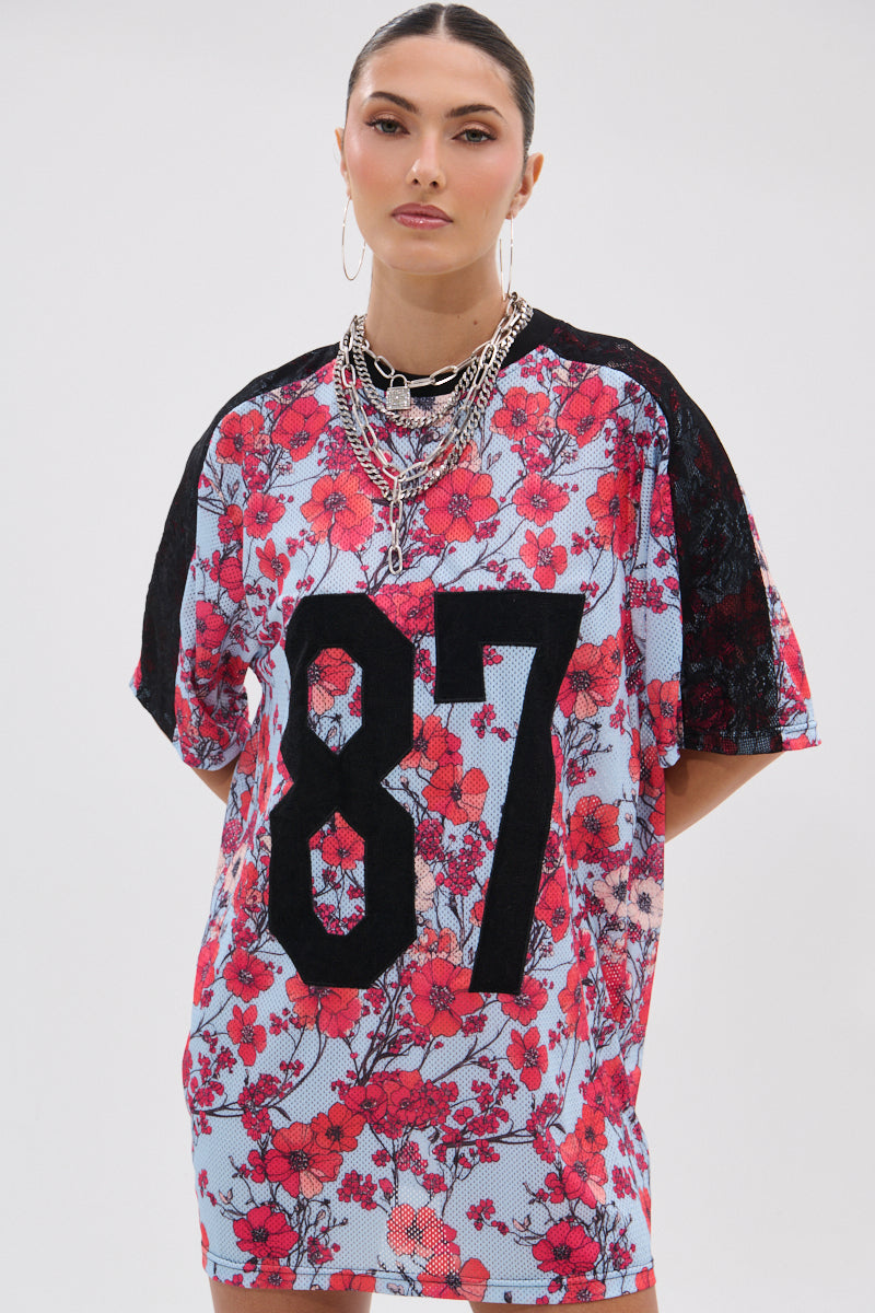 A woman stands against a plain white background wearing the POPPIN POPPIES LACE JERSEY DRESS, featuring a loose poppy print and bold black "87" on the front. She accessorizes with silver necklaces, hoop earrings, and has her hair pulled back. 3