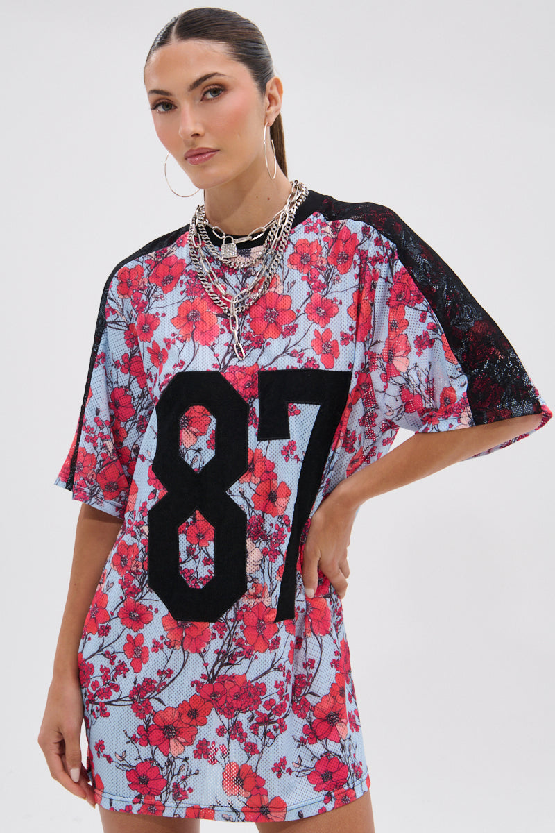 A woman wears the POPPIN POPPIES LACE JERSEY DRESS, featuring a loose fit, red floral pattern, black lace sleeves, and a bold black "87" on the front. She completes her look with layered necklaces and hoop earrings for added femininity. 4