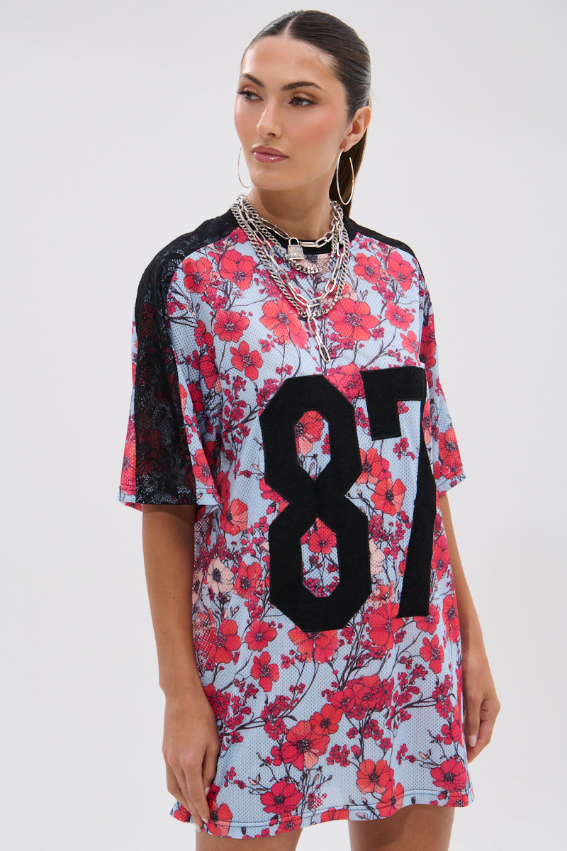 A woman models the POPPIN POPPIES LACE JERSEY DRESS, featuring a bold “87”, floral lace details, and black sleeves, styled with layered silver necklaces and hoop earrings against a plain white background. 5
