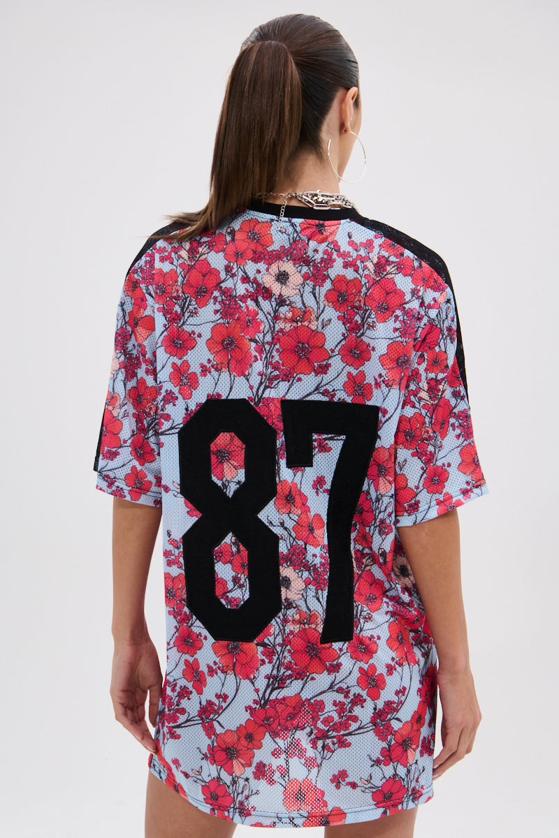 A person with long brown hair in a ponytail models the POPPIN POPPIES LACE JERSEY DRESS, featuring a loose mesh design, red floral pattern, and large black "87" on the back, against a plain white background. 7