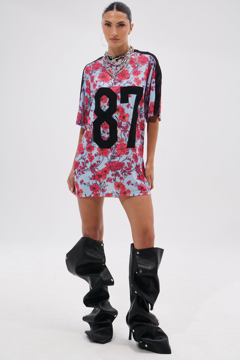 A woman stands confidently in the POPPIN POPPIES LACE JERSEY DRESS—a loose floral piece with a bold black “87” on the front—styled with layered necklaces, hoop earrings, and oversized black slouchy over-the-knee boots against a plain white background. 8