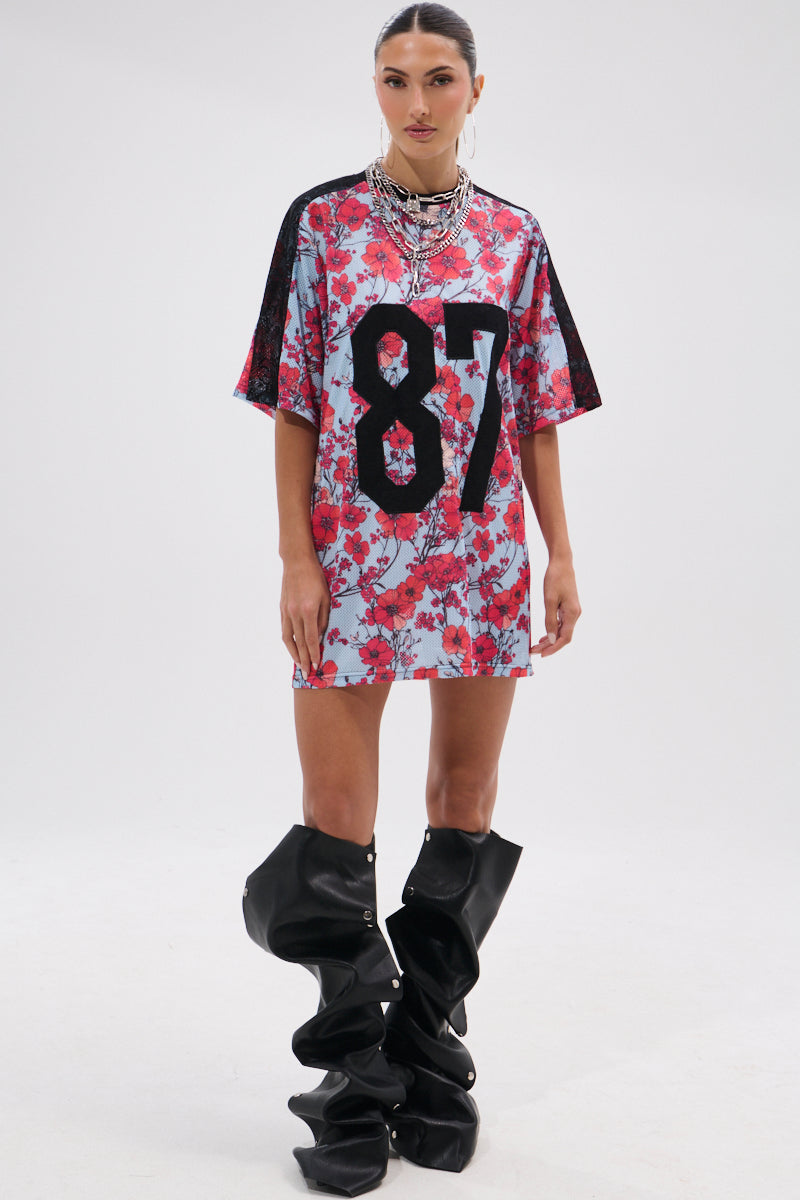 A woman poses against a plain background in the POPPIN POPPIES LACE JERSEY DRESS, a loose floral piece with the number 87 in black on the front. She completes the look with oversized slouchy black boots and layered silver necklaces. 9