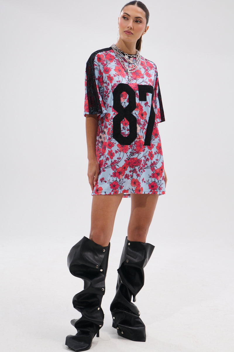 A woman wearing the POPPIN POPPIES LACE JERSEY DRESS, featuring a loose floral design and bold black "87" on the front, with chunky layered necklaces and slouchy black knee-high boots, stands against a plain white background. 10