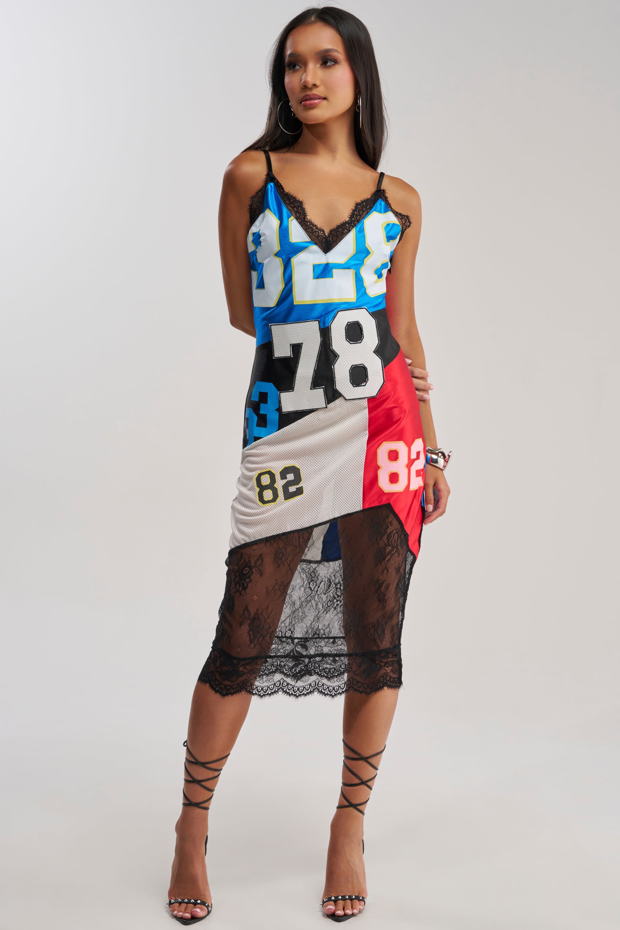 A woman in the SPORT CHIC PANELED LACE MIDI DRESS, featuring colorful panels with bold numbers and lace hem, pairs it with black strappy heels against a plain background.