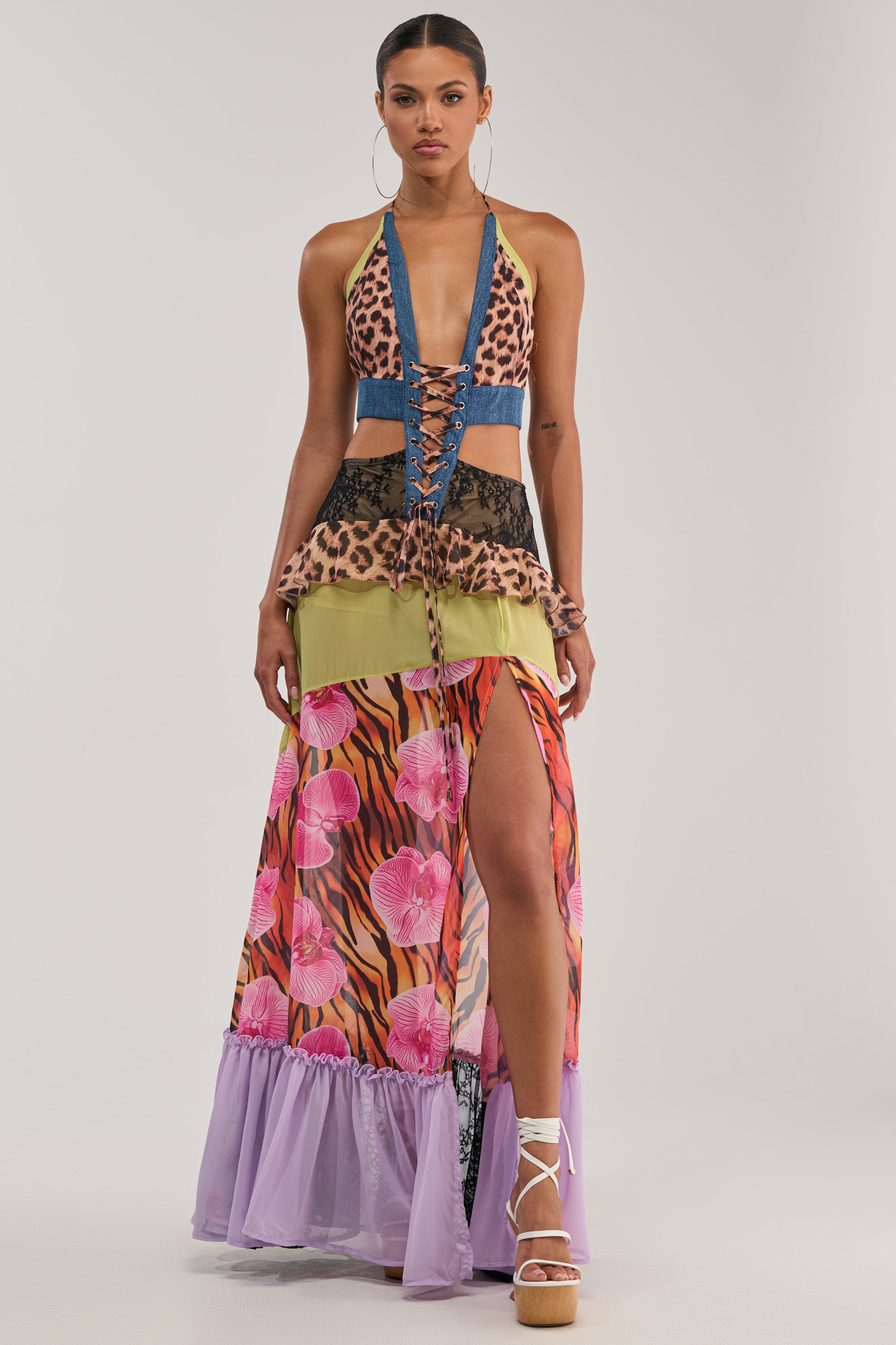 A woman wears the SUPER SWEET GIRL PATTERNED MAXI DRESS—a bold statement piece with a leopard print halter top, lace-up front, mixed patterns, green and leopard skirt layers, pink floral accents, lavender ruffle hem—plus hoop earrings and platform sandals.