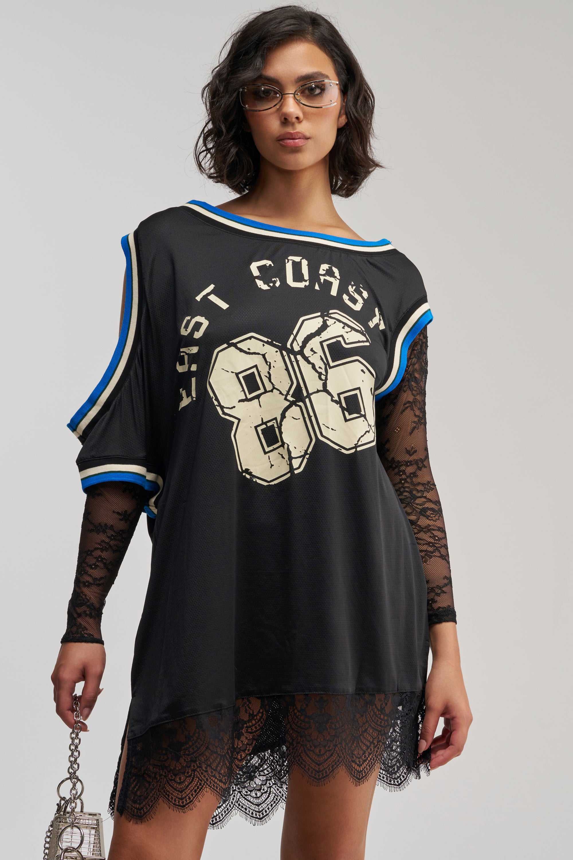 A woman wearing sunglasses and the PUT ME IN COACH LACE MINI JERSEY DRESS—featuring a loose black fit, bold “East Coast 86” lettering, lace sleeves, and hem—holds a small handbag while posing confidently against a plain background. This sporty-chic dress is perfect for creating a standout streetwear look and making a bold statement.