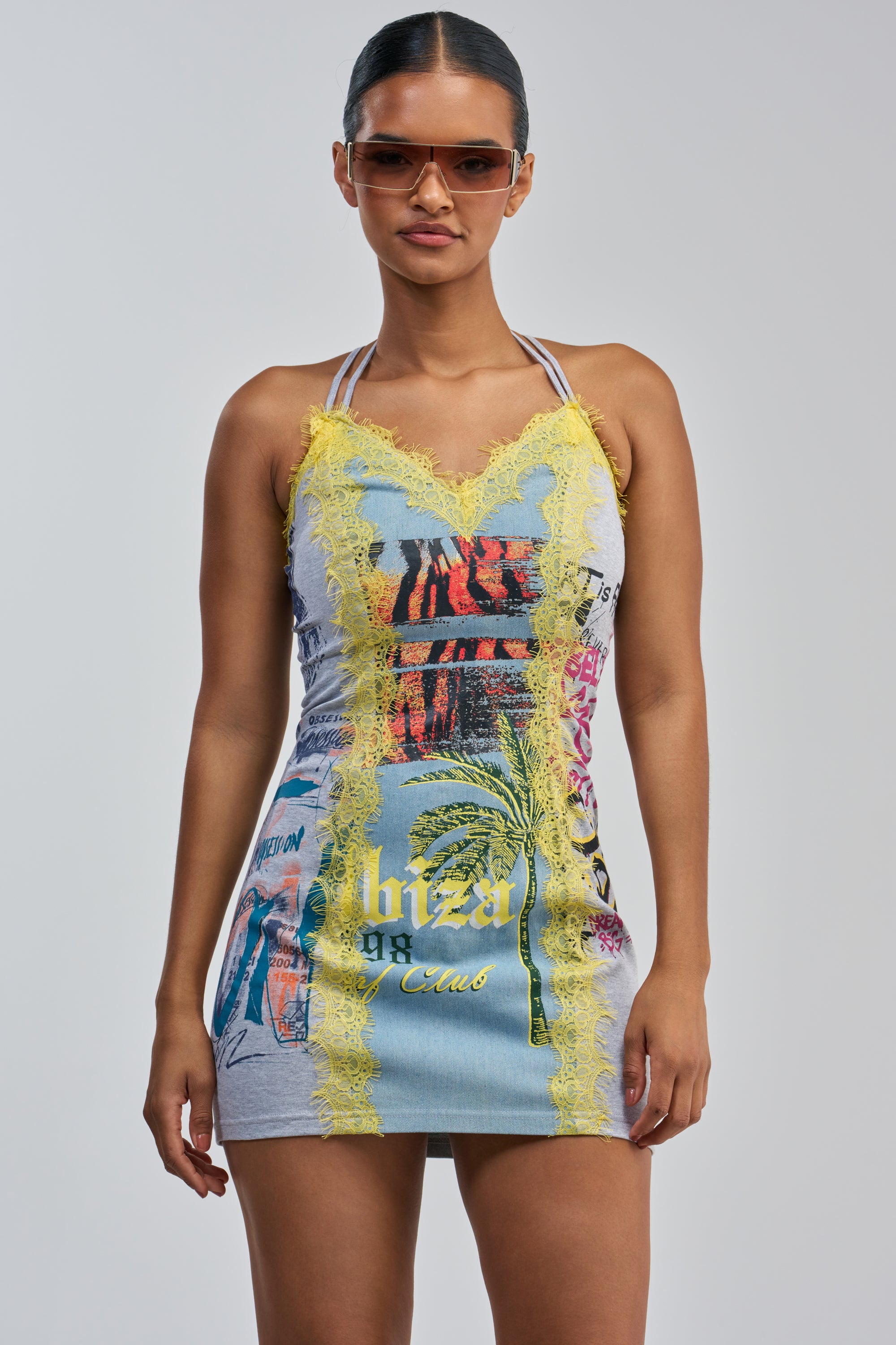 A woman stands against a light gray background wearing the JUST BETWEEN US MINI DRESS YELLOW MULTI— a sleeveless, fitted streetwear mini dress featuring multicolored graphics and yellow lace trim, paired with sunglasses.