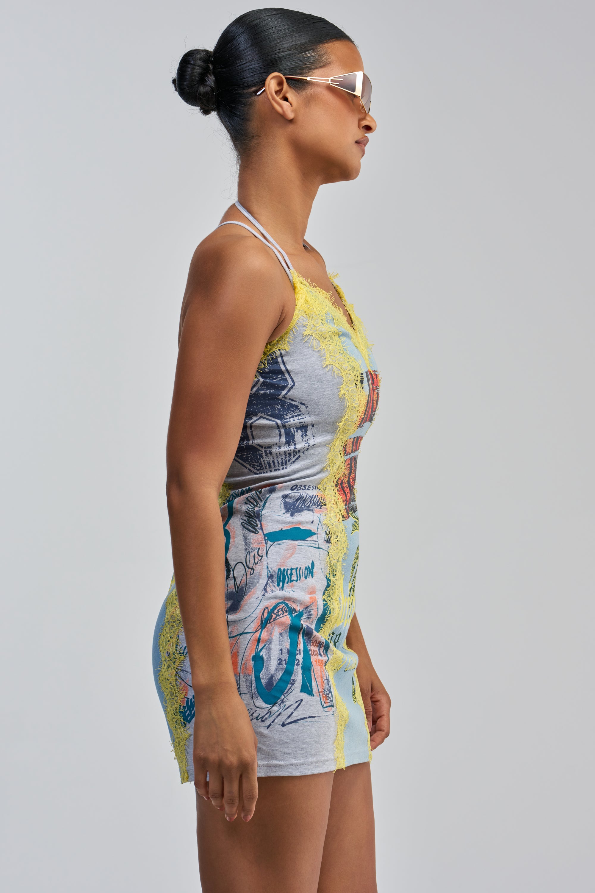 A woman with dark hair in a bun and sunglasses is shown in side profile wearing the JUST BETWEEN US MINI DRESS YELLOW MULTI—a sleeveless streetwear mini dress featuring a graphic print and yellow lace trim—against a plain light gray background.