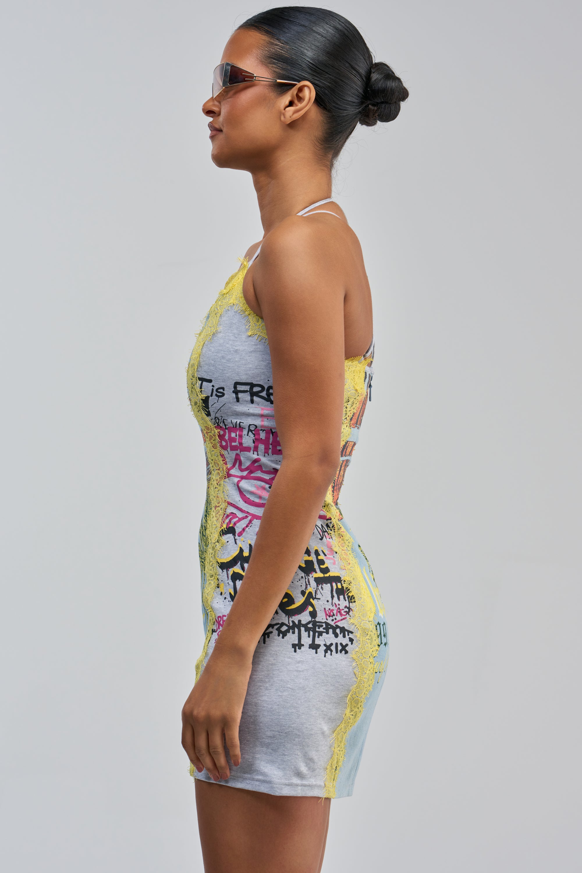A woman in sunglasses, wearing the JUST BETWEEN US MINI DRESS YELLOW MULTI—a gray halter-style streetwear mini dress featuring yellow lace trim and colorful graffiti-inspired designs—stands in profile against a plain background.