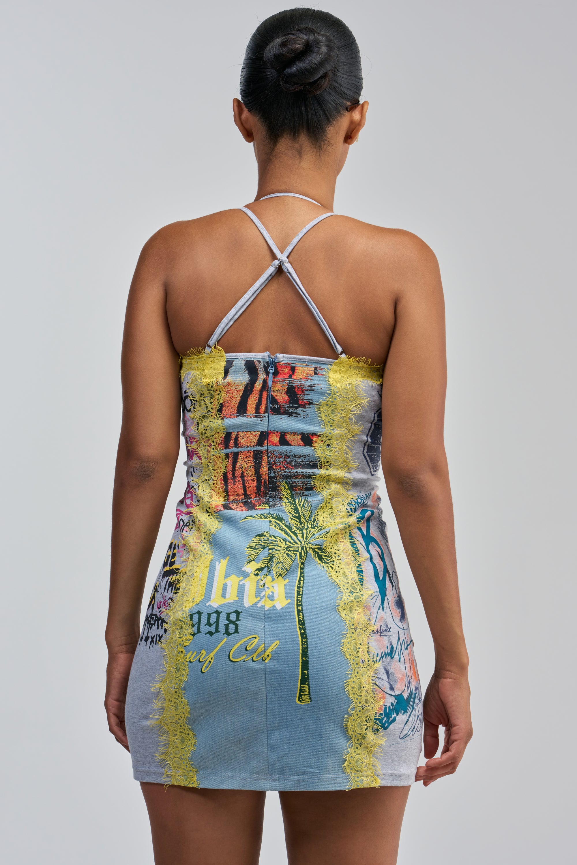 A woman stands facing away, wearing the JUST BETWEEN US MINI DRESS YELLOW MULTI— a streetwear mini dress featuring vibrant graphic prints and yellow lace trim. Her hair is styled in a neat bun against a plain background.