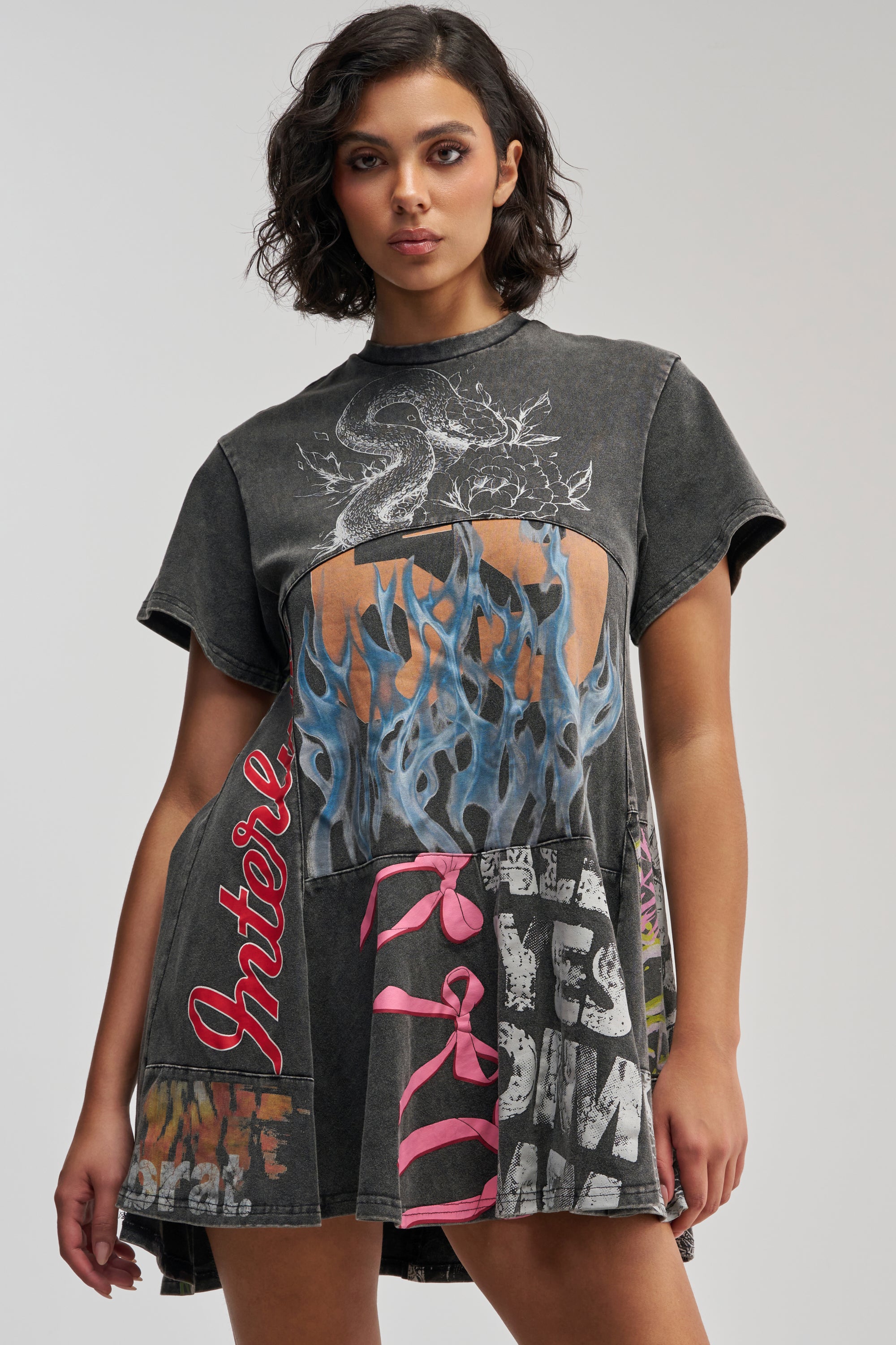 A woman with wavy dark hair wears the COLD HEARTED GRAPHIC MINI DRESS IN BLACK MULTI, a grunge-inspired streetwear mini dress with short sleeves and bold graphic designs—including a dragon, blue flames, pink bow images, graffiti-style text, and the word “Interior” in red on one side. She stands against a plain background.