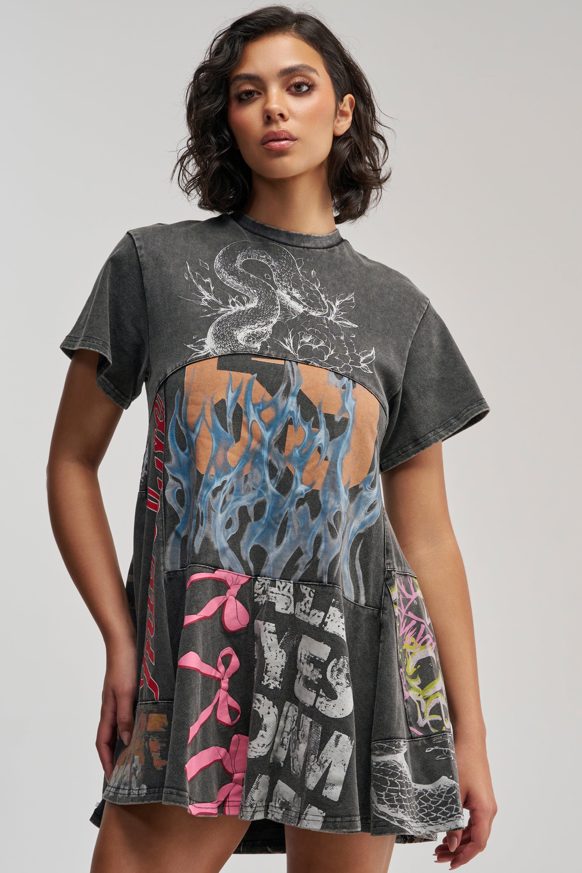 A woman wearing the COLD HEARTED GRAPHIC MINI DRESS IN BLACK MULTI—a short-sleeve, grunge-inspired patchwork dress featuring dragon and flames graphic prints with striking pink accents—stands against a plain background, looking confidently at the camera.