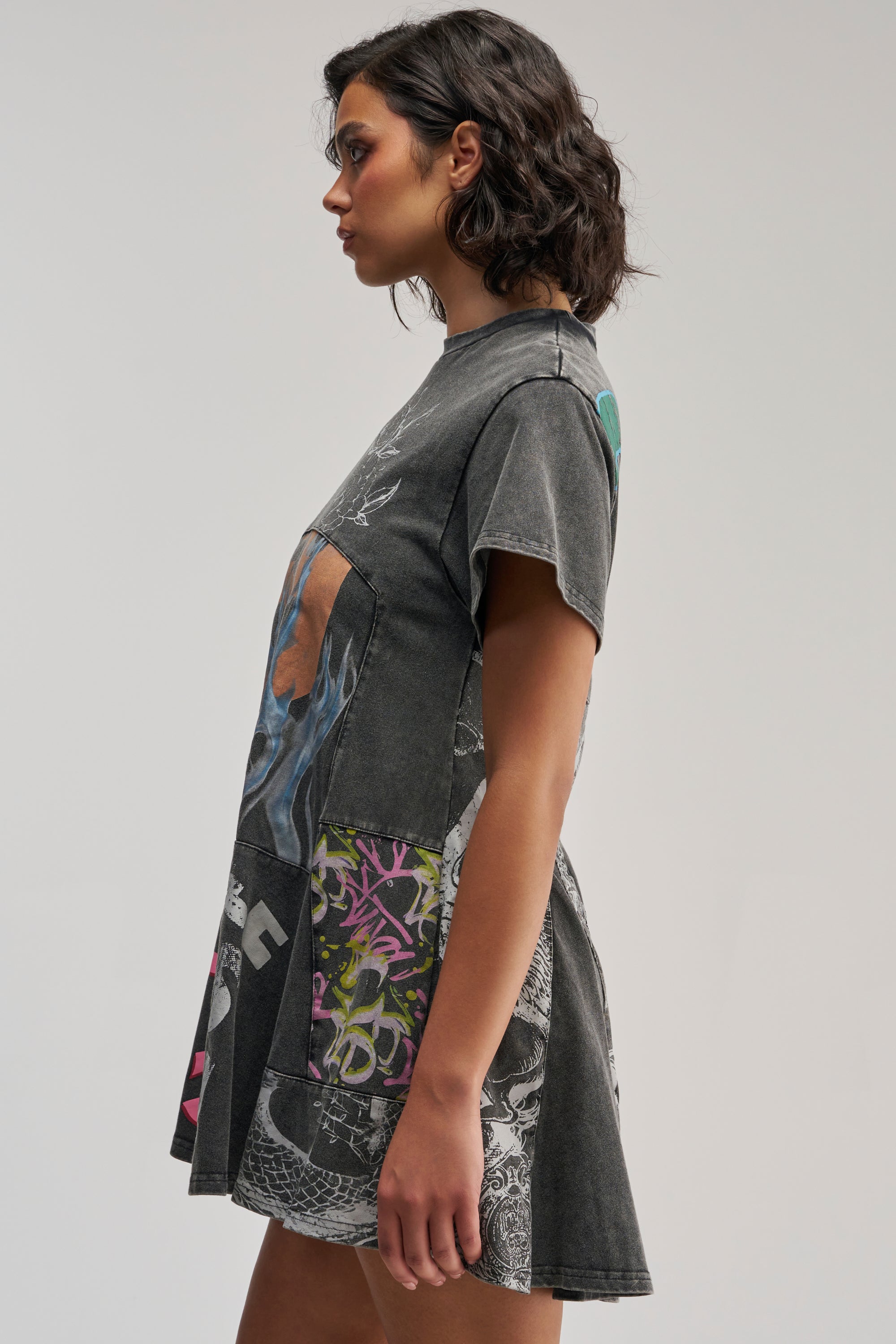A woman with short, wavy dark hair stands in profile wearing the COLD HEARTED GRAPHIC MINI DRESS IN BLACK MULTI, which features colorful and grayscale designs, against a plain background.