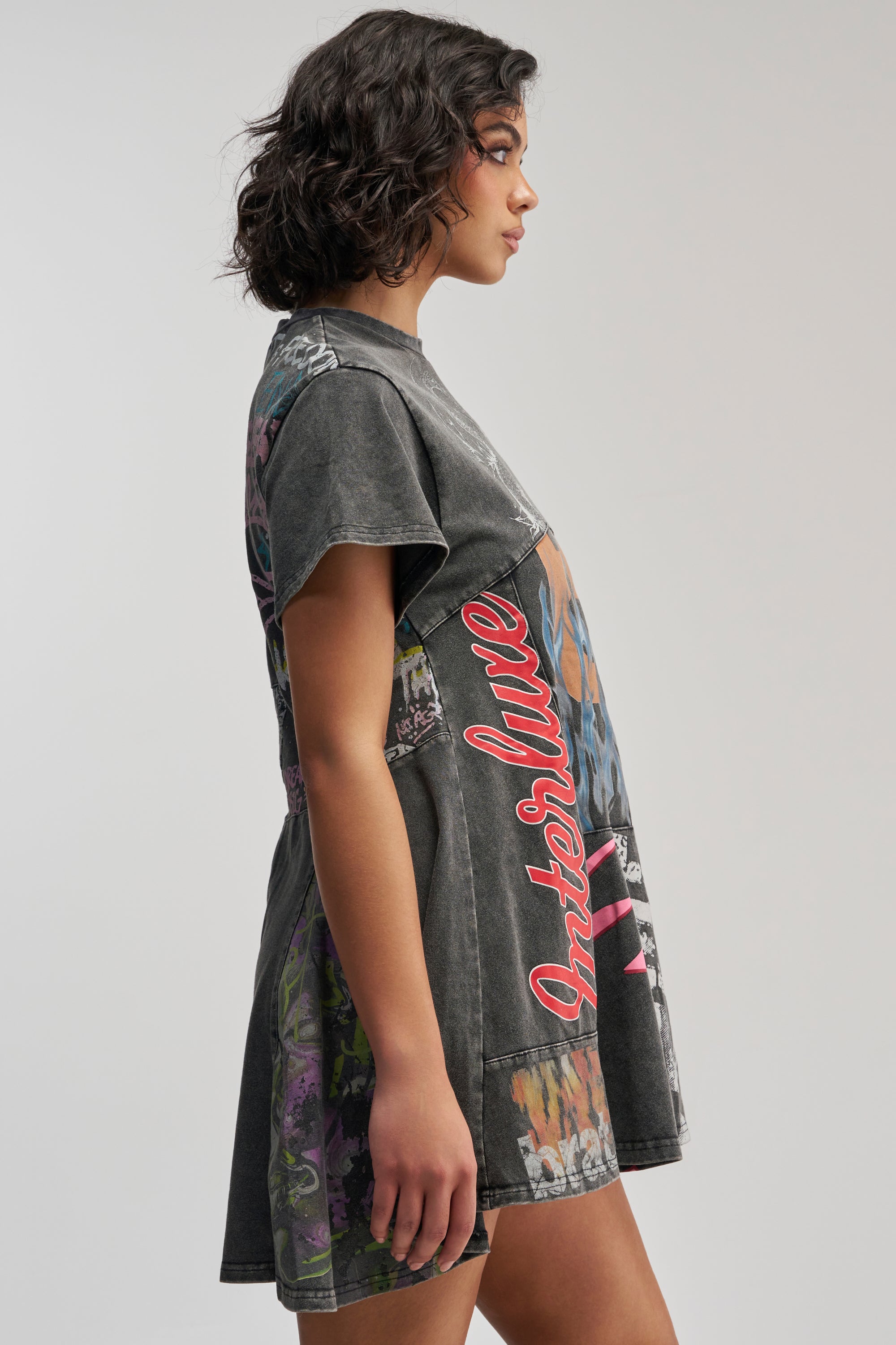 A woman with wavy dark hair is shown in profile against a plain light background, wearing the COLD HEARTED Graphic Mini Dress in Black Multi—a short-sleeved, grunge-inspired dress featuring colorful patterns and the word “Interlude” in red.