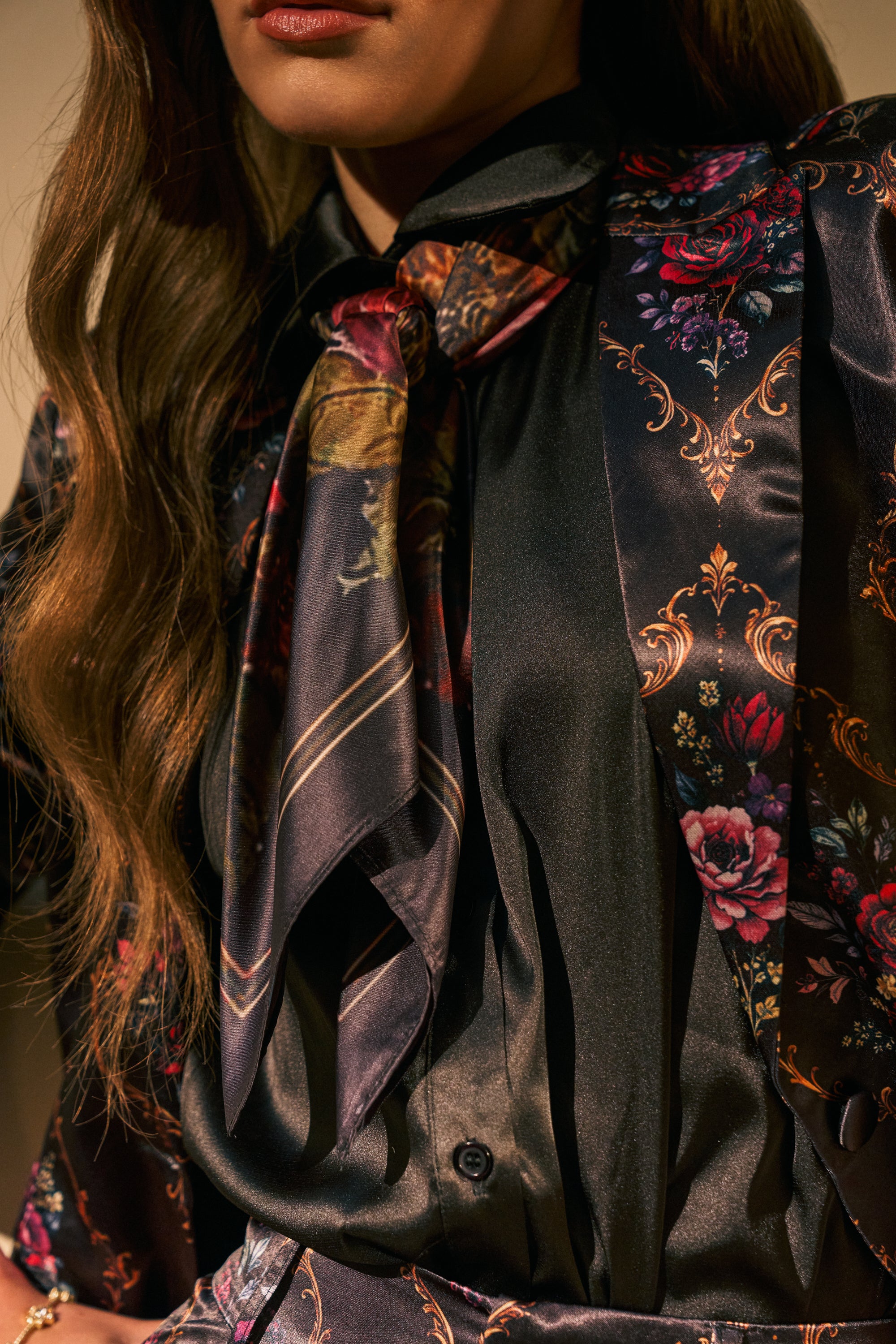 A person with wavy brown hair wears a silky black blouse adorned with colorful floral and ornate patterns, accessorized with the MIDNIGHT GARDEN SILK SCARF tied in a loose bow at the neck.