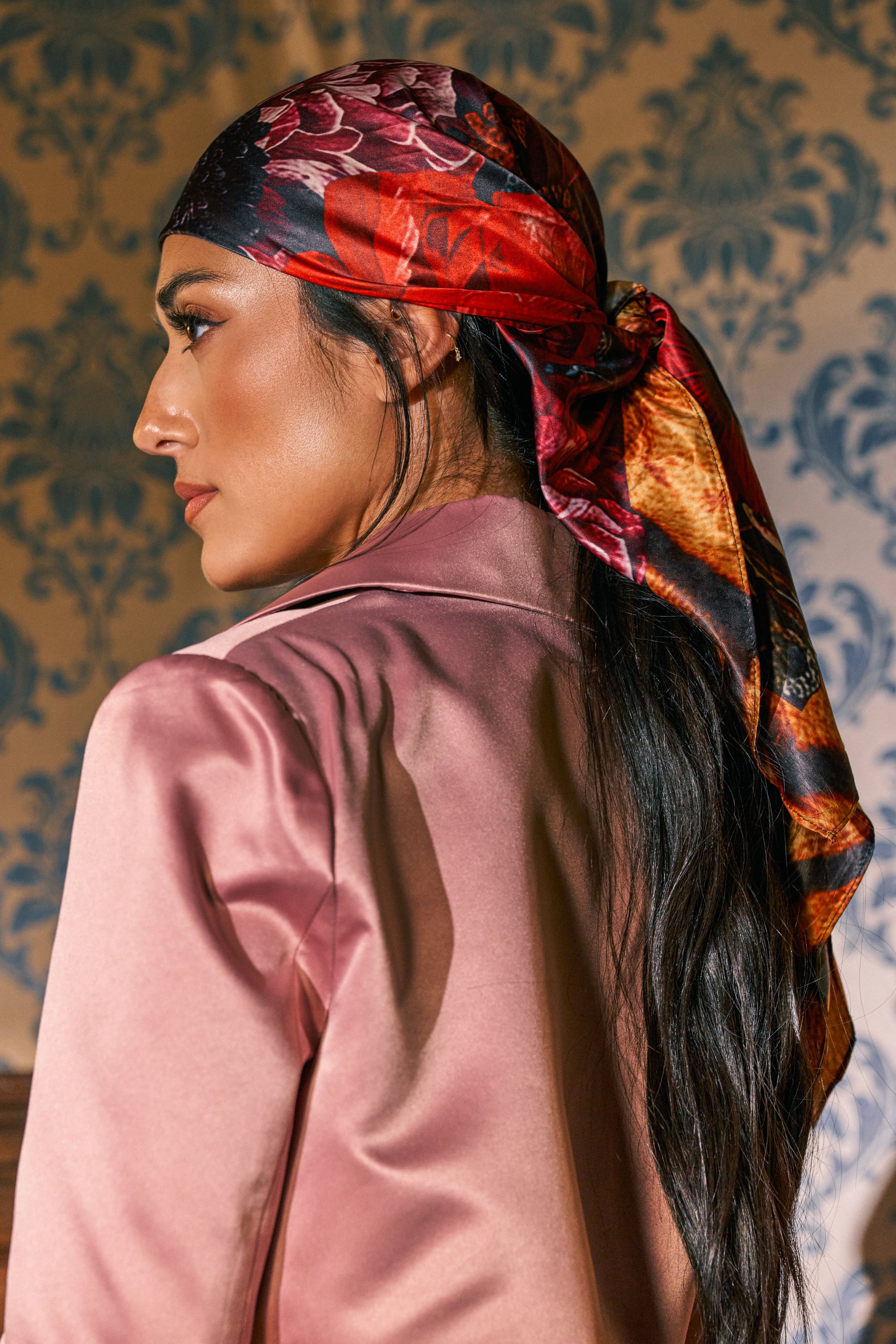 Wearing the UNTAMED PETALS SCARF as a chic hair accessory, a woman with long dark hair and a silky pink blazer stands before patterned wallpaper, her profile visible as she faces away.