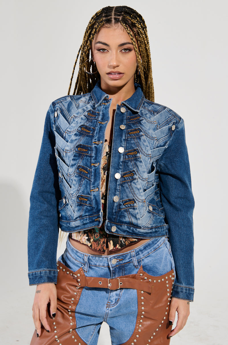 A person with long braided hair wears the BOTTOMS UP DENIM JACKET, known for its zigzag design, paired with brown leather pants. They stand against a simple background, creating a striking cropped silhouette.