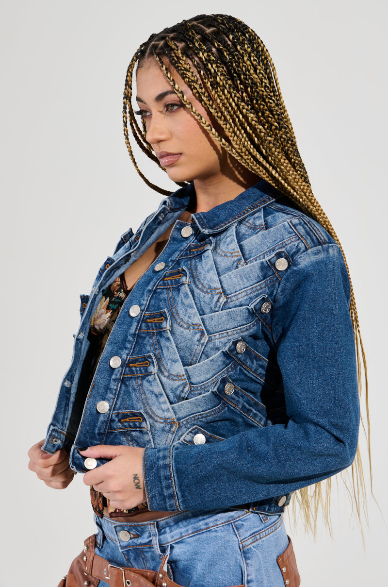 A person with long braided hair wears the BOTTOMS UP DENIM JACKET, showcasing its zigzag design and button details. They hold the jacket collar, unveiling a floral top beneath it. The outfit is completed with denim pants and a brown belt, all set against a plain light gray background.