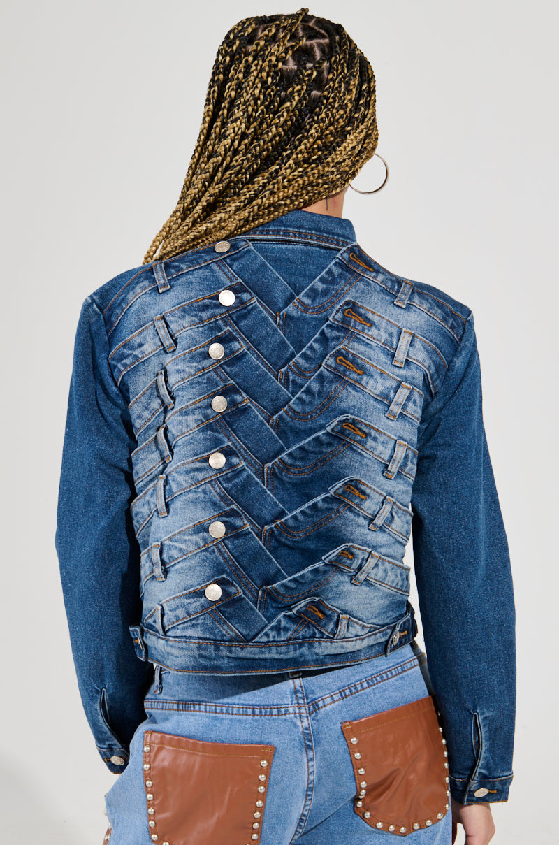 The image displays a person from behind wearing the BOTTOMS UP DENIM JACKET, distinguished by its intricate zigzag design on the back and adorned with silver buttons. The individual, who has braided hair, is also sporting jeans that feature leather pocket accents and metal studs.
