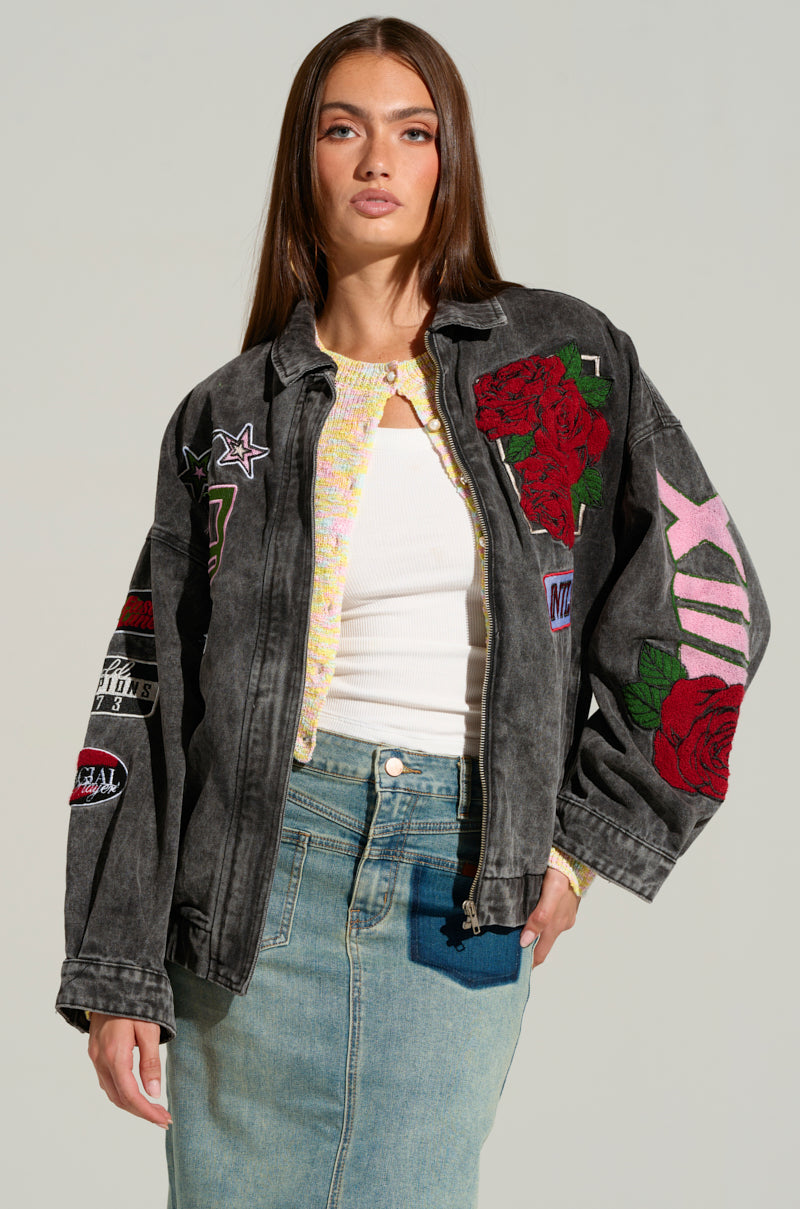 A woman with long brown hair stands against a plain background, showcasing the ROSES ARE RED EMBROIDERED DENIM BOMBER. The dark grey denim jacket is adorned with various racing-inspired embroidered patches and floral designs, including red roses and numbers. She complements her look with a white top and a light blue denim skirt, gazing confidently at the camera.
