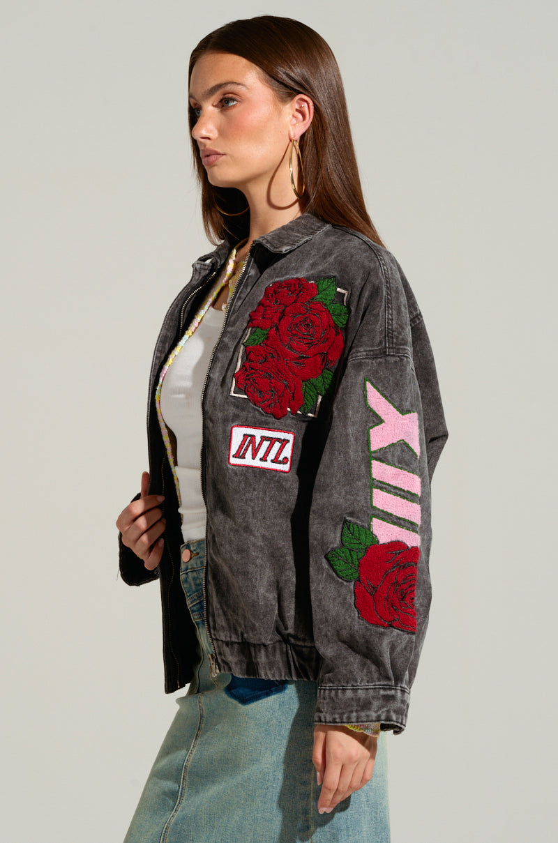A young woman with long brown hair wears the "ROSES ARE RED EMBROIDERED DENIM BOMBER," a distressed black jacket featuring racing-inspired embroidered patches and red rose designs. Large hoop earrings complement her light-colored top, paired with high-waisted blue jeans. She faces left, gazing forward with a bold look.