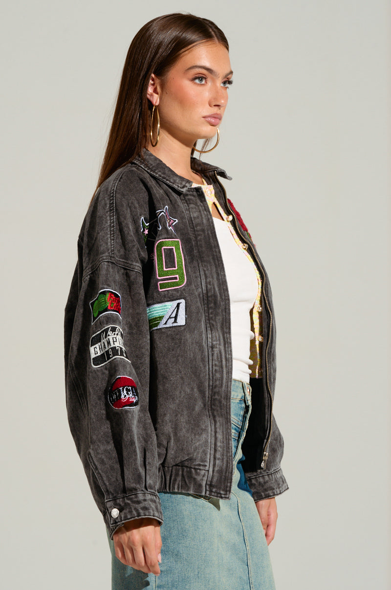 A woman with long brown hair dons the ROSES ARE RED EMBROIDERED DENIM BOMBER, a black denim jacket adorned with racing-inspired embroidered patches. She pairs it with a white top and blue denim skirt, while large hoop earrings dangle as she gazes left. The background is plain.