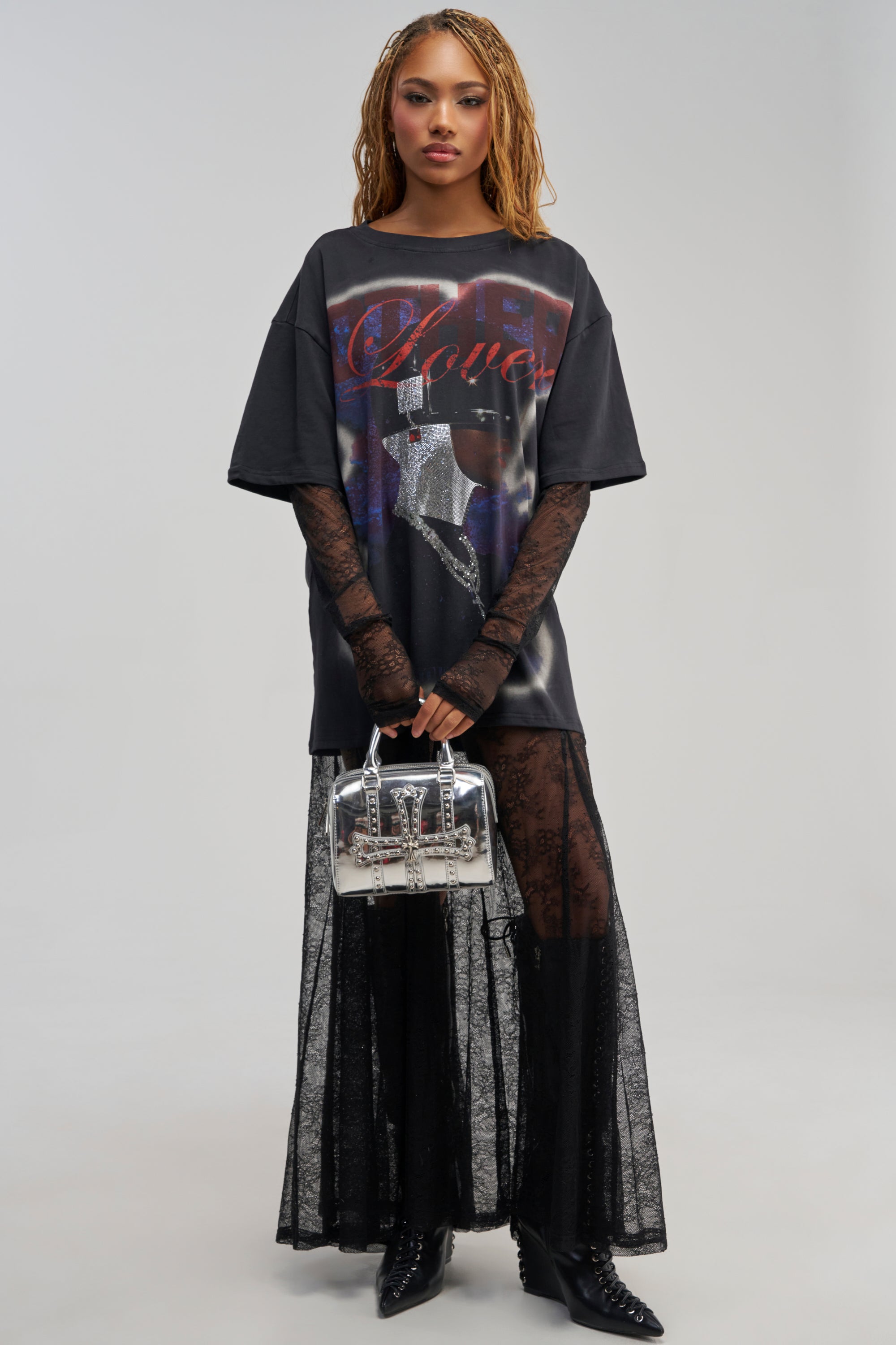 A woman wears the GIRLY BUT NOT LAYERED LACE MIDI DRESS IN BLACK MULTI, styled with black boots and a small silver handbag for an edgy streetwear-inspired look. The light background highlights the dress’s unique graphic details, sheer lace sleeves, and layered black lace skirt.