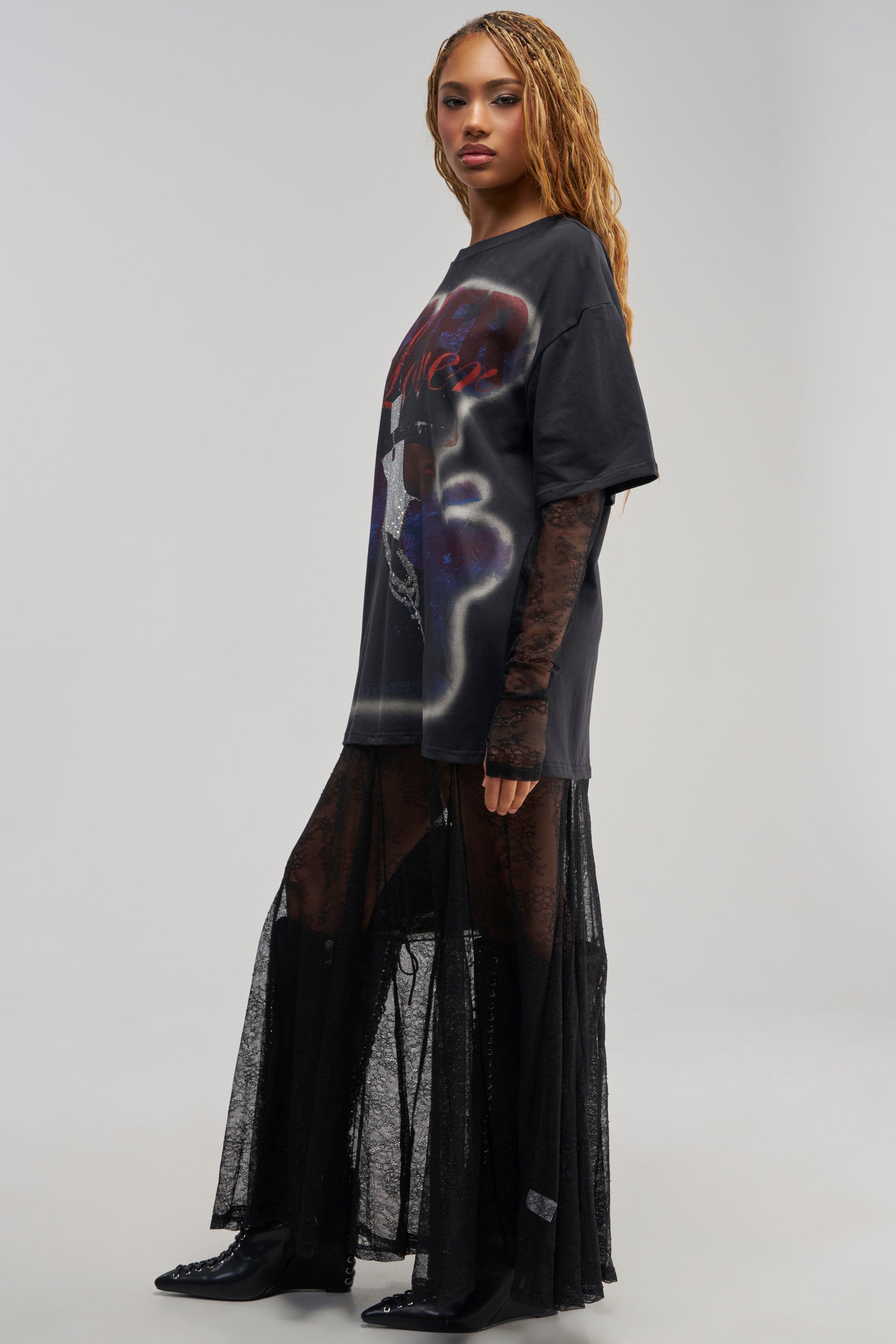 A woman stands sideways against a plain light background, wearing the GIRLY BUT NOT LAYERED LACE MIDI DRESS IN BLACK MULTI. The dress features an oversized streetwear-inspired graphic t-shirt layered over a sheer black lace midi skirt. She has long wavy hair and black shoes.