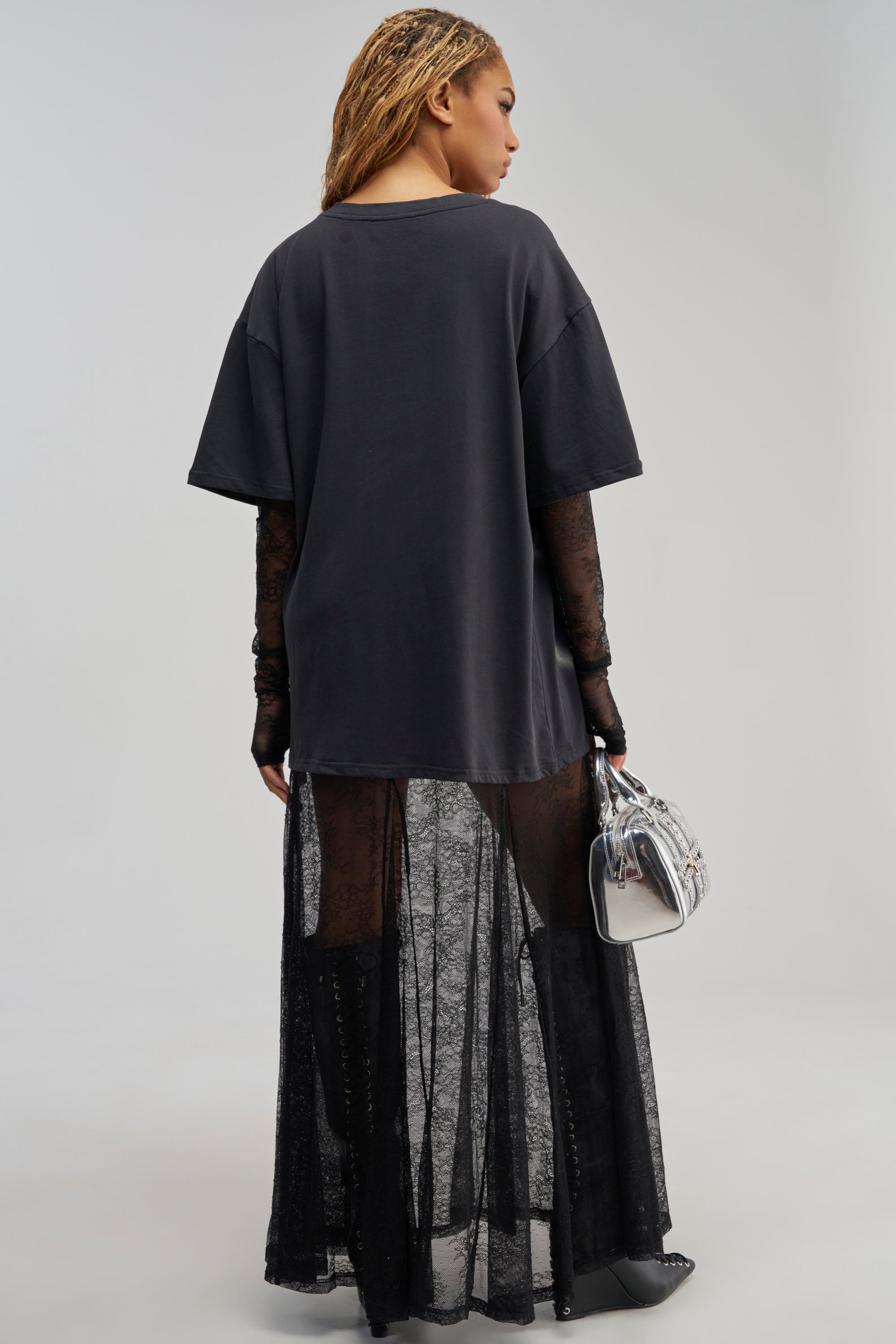 Model faces away, wearing the GIRLY BUT NOT LAYERED LACE MIDI DRESS IN BLACK MULTI, styled with lace gloves and a shiny silver handbag. The layered lace and relaxed fit of the dress create an edgy streetwear look, set against a plain light gray background.