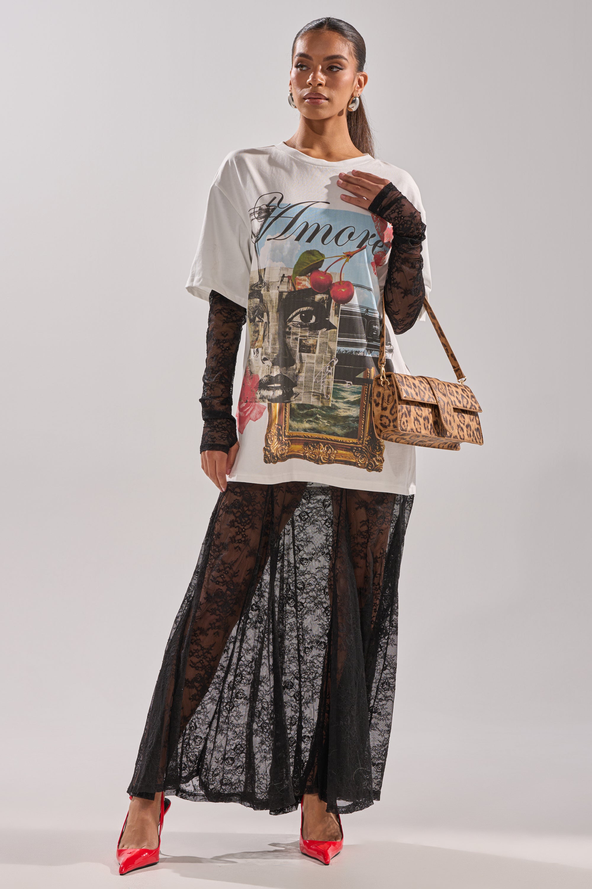 A woman models the GIRLY BUT NOT LAYERED LACE MIDI DRESS WITH GRAPHIC, styled with black lace gloves, red heels, and a tan patterned shoulder bag against a neutral background.