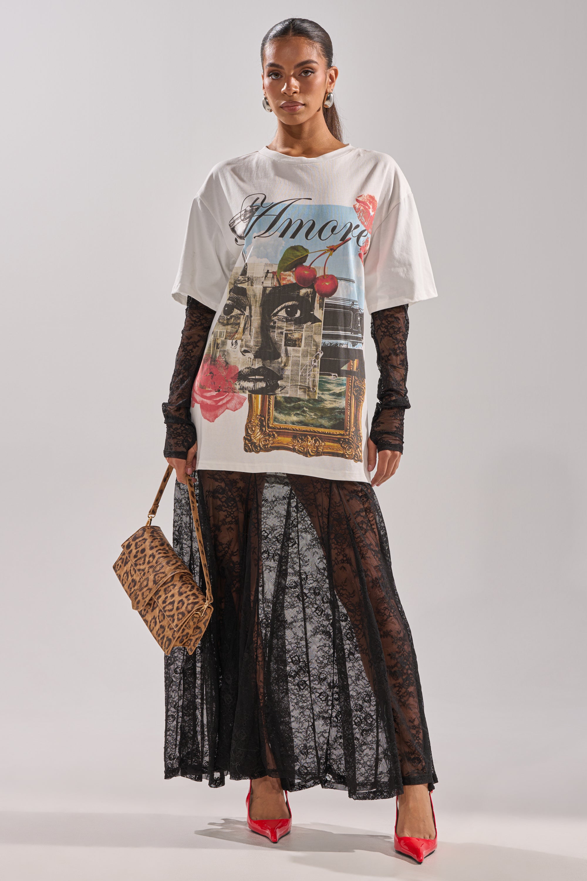 A woman wears the GIRLY BUT NOT LAYERED LACE MIDI DRESS WITH GRAPHIC, styled over a sheer black lace layer. She carries a leopard print handbag, wears red heels and hoop earrings, and stands against a plain light background with her hair pulled back.