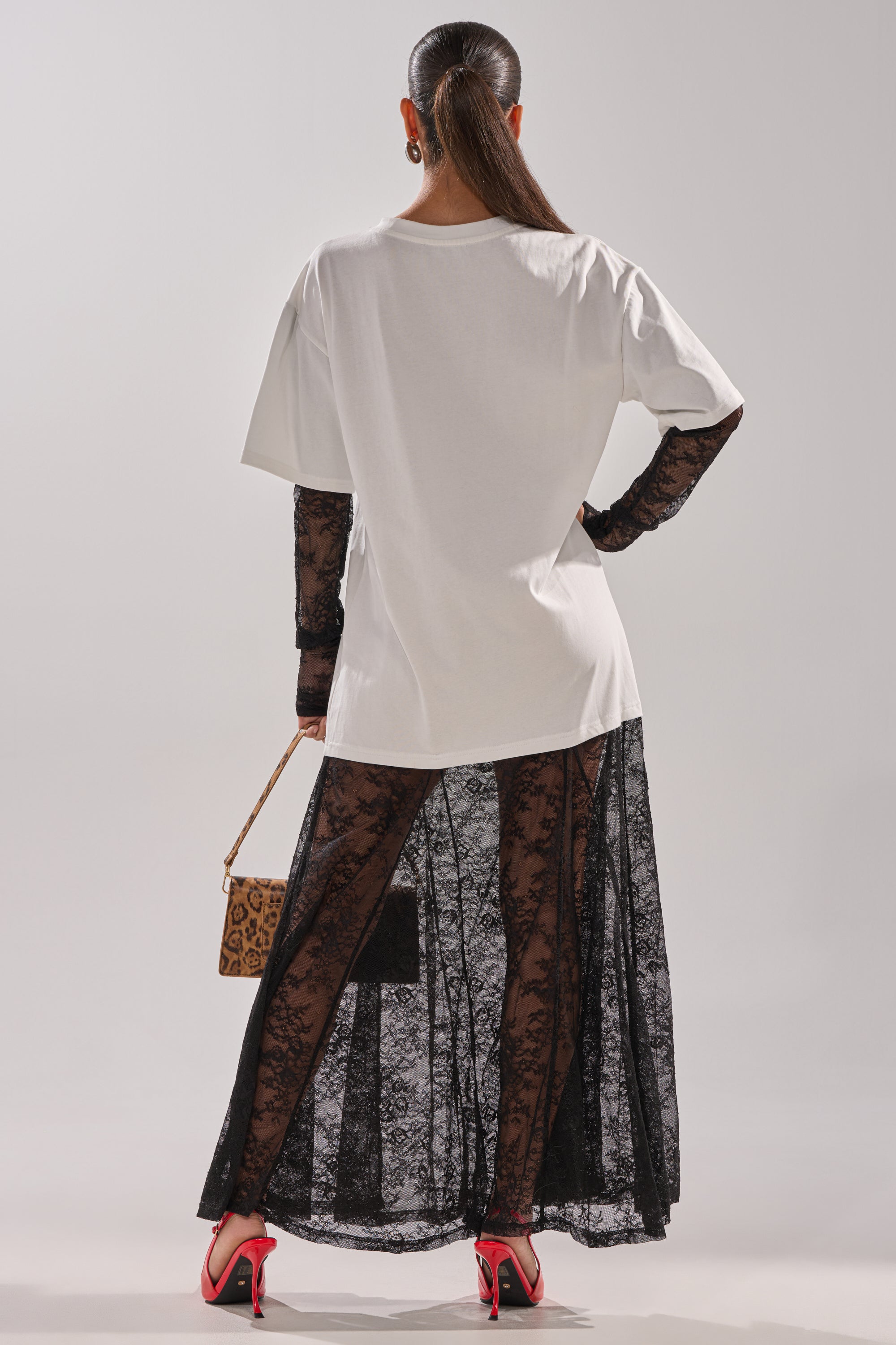 A woman faces away, wearing the GIRLY BUT NOT LAYERED LACE MIDI DRESS WITH GRAPHIC styled with a white oversized T-shirt, animal-print purse, bright red heels, and a ponytail.