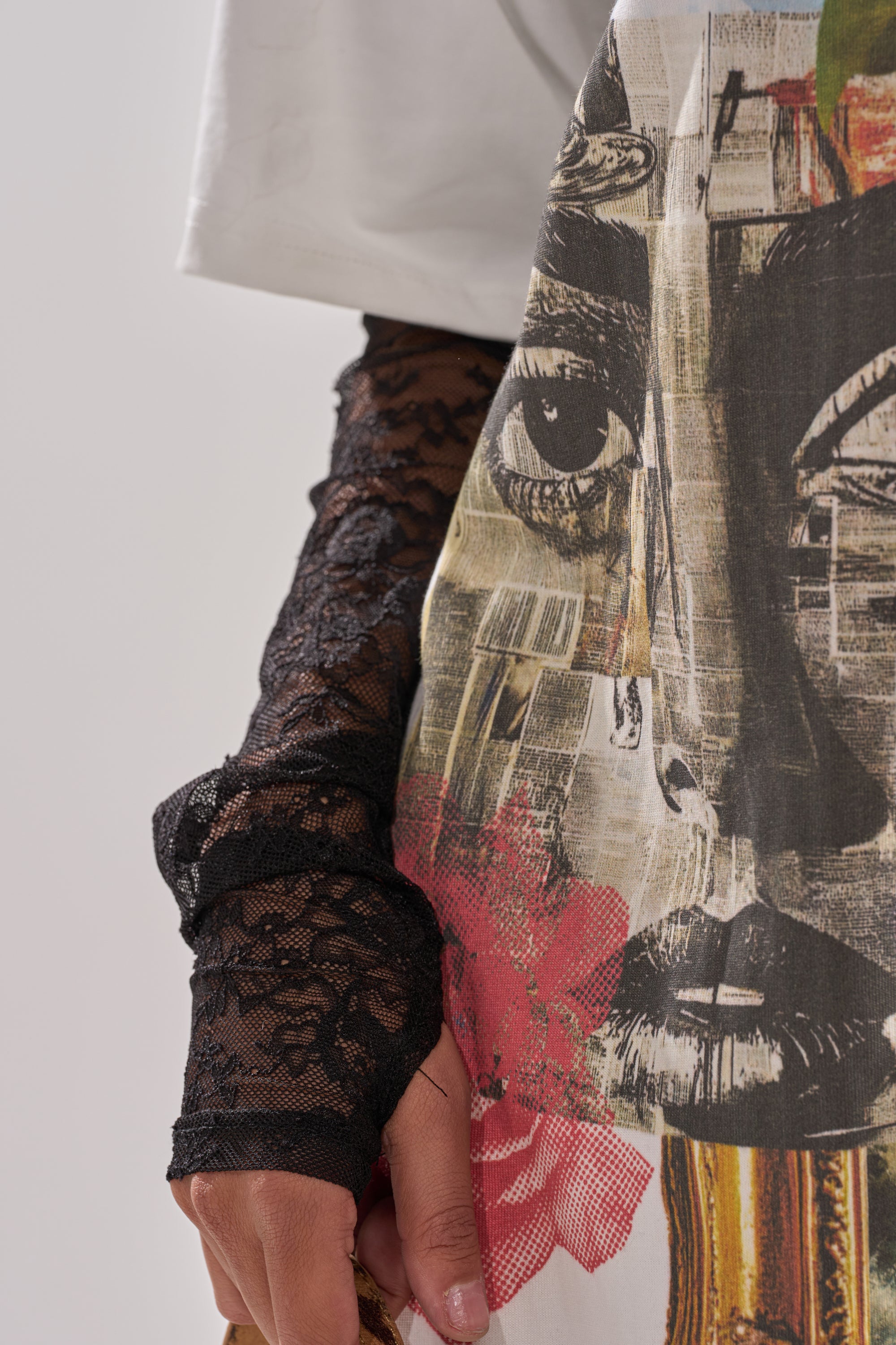 A person wears the GIRLY BUT NOT LAYERED LACE MIDI DRESS WITH GRAPHIC, featuring an abstract face print with red and gold accents, layered over a white shirt and sheer black lace gloves. Only the lower face, hand, and skirt design are visible.