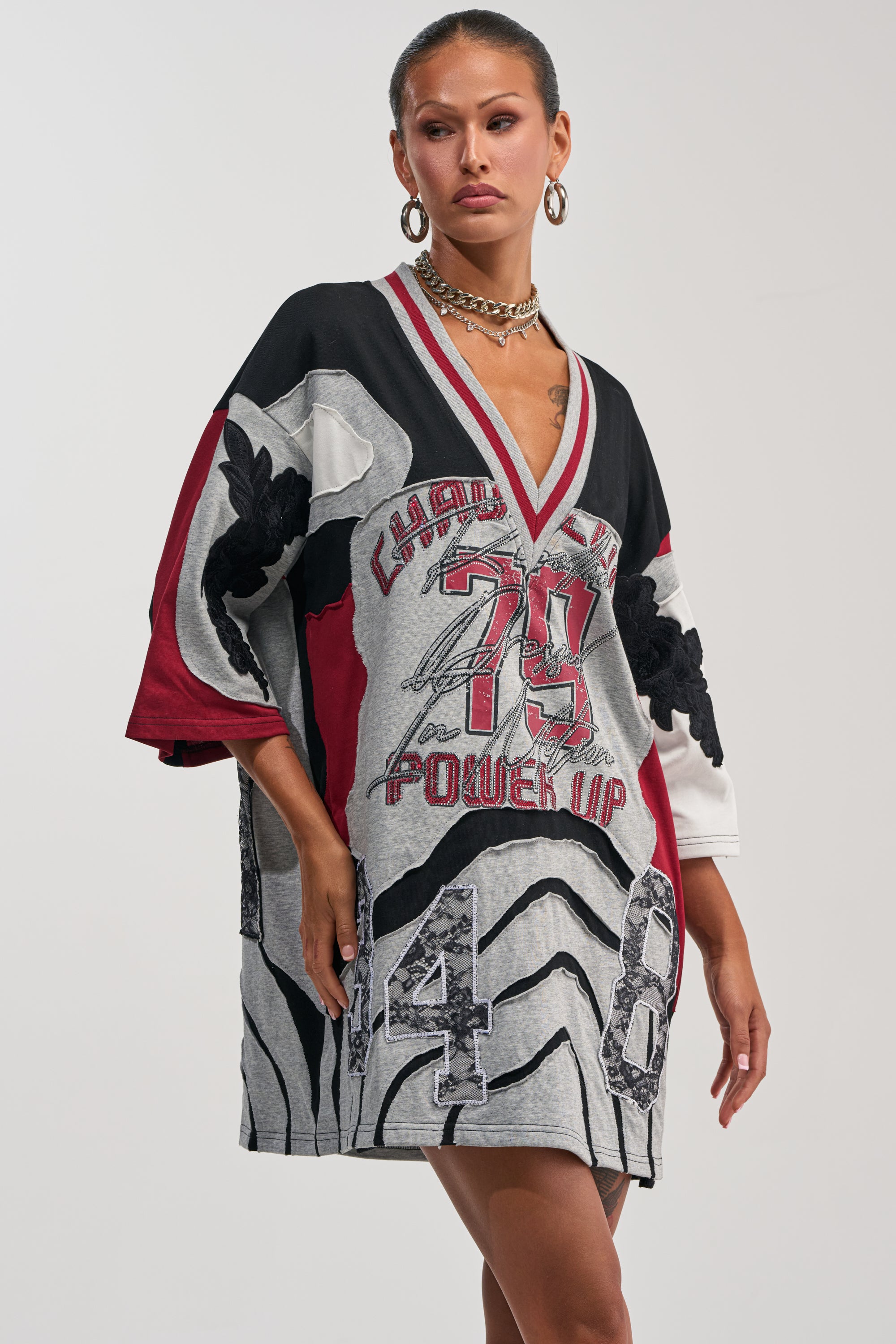 A woman wears the AFTER HOURS GRAPHIC MINI DRESS, an oversized varsity-inspired piece featuring bold patterns, numbers, and text in red, black, gray, and white. She completes her look with large hoop earrings and layered necklaces.