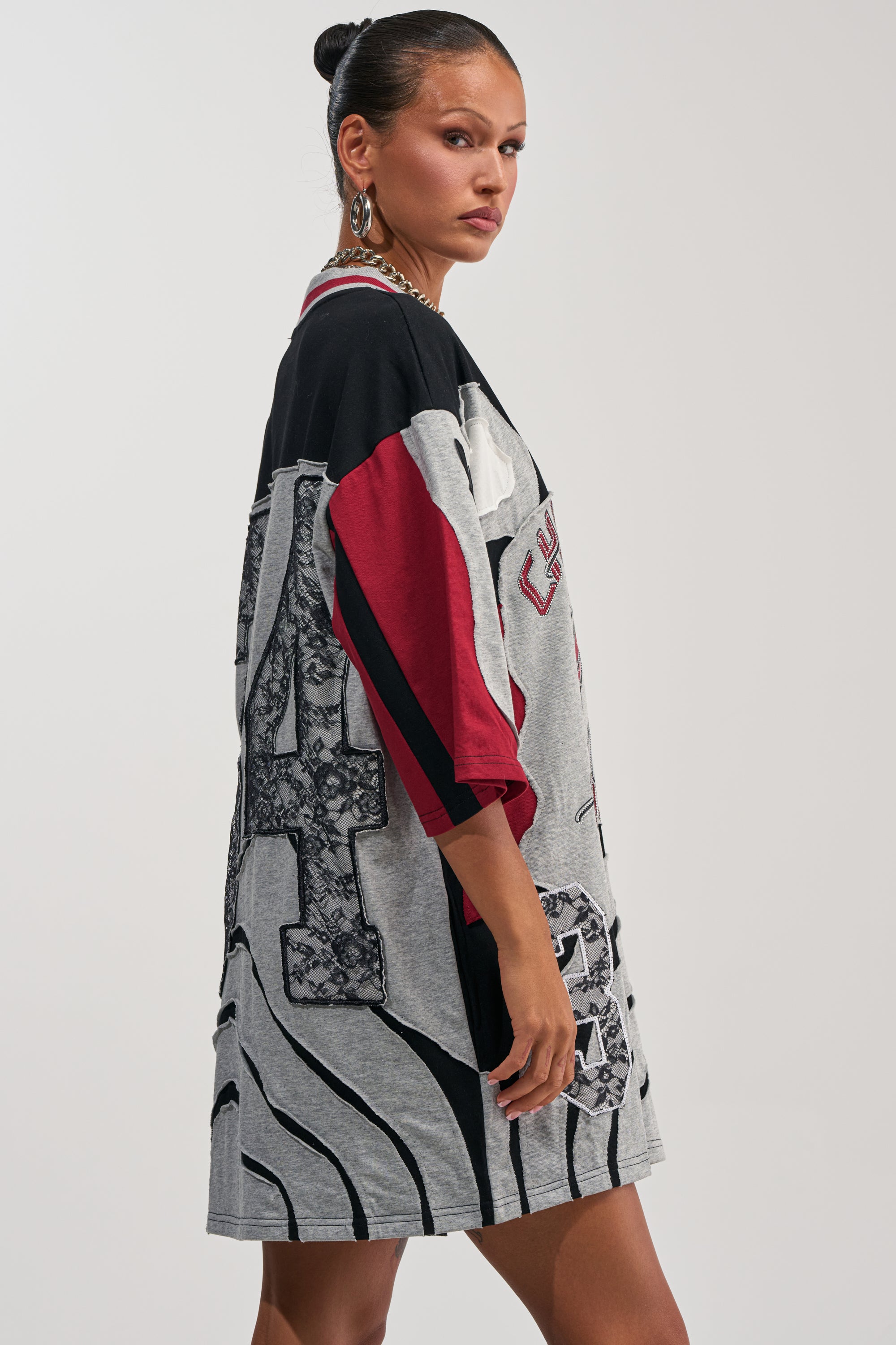 A woman with dark hair in a bun wears the AFTER HOURS GRAPHIC MINI DRESS—an oversized mini dress featuring bold color-blocked patterns in gray, black, and red—as she stands sideways and looks over her shoulder against a plain background.