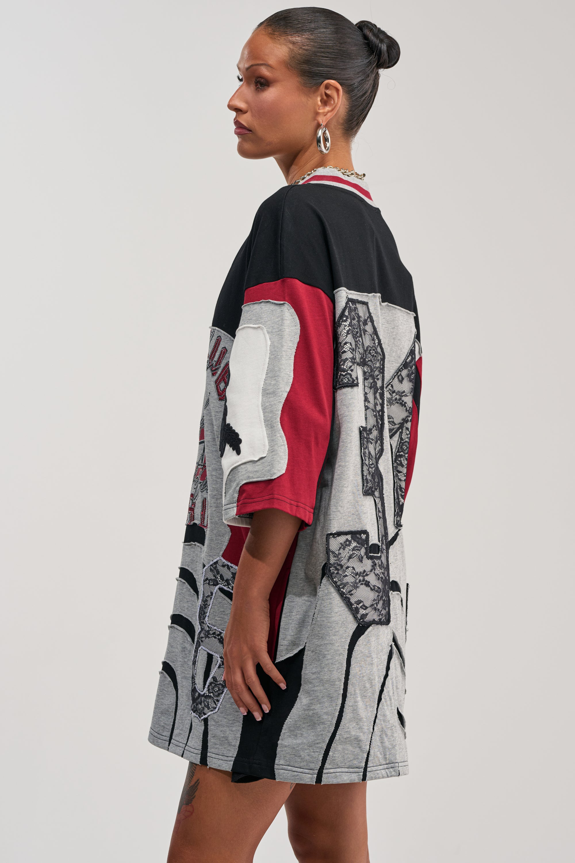 A woman stands sideways wearing the AFTER HOURS GRAPHIC MINI DRESS—an oversized patchwork tee mini dress with black, gray, and red graphic prints. Her hair is styled in a bun, and she accessorizes with earrings and a necklace.