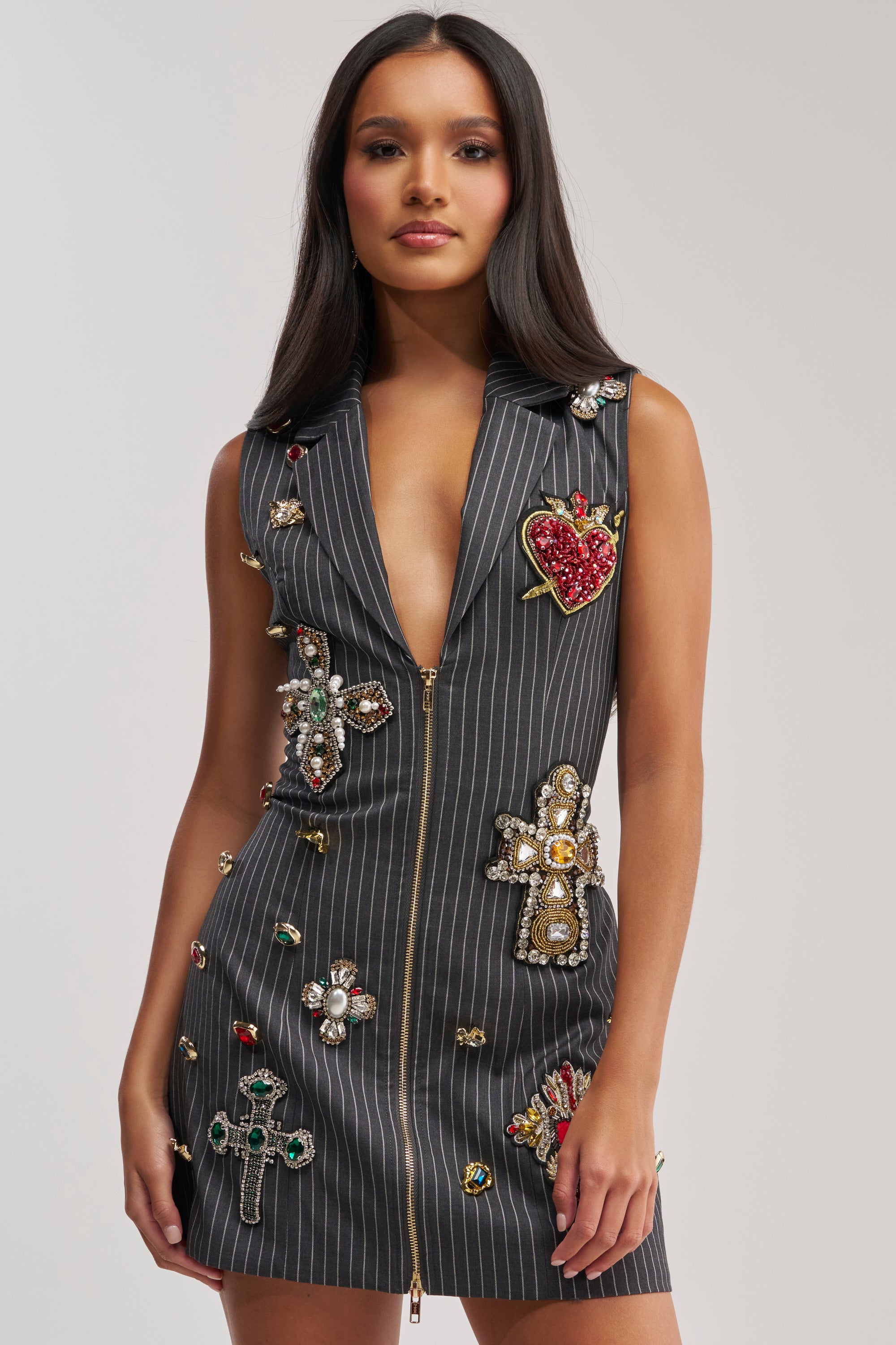 Wearing the PRETTIEST PROBLEM SLEEVELESS PATCHWORK MINI DRESS—a grey pinstripe mini with jeweled appliqué crosses, hearts, and gems—a woman stands against a plain background, embodying the look of a gemstone accent dress.