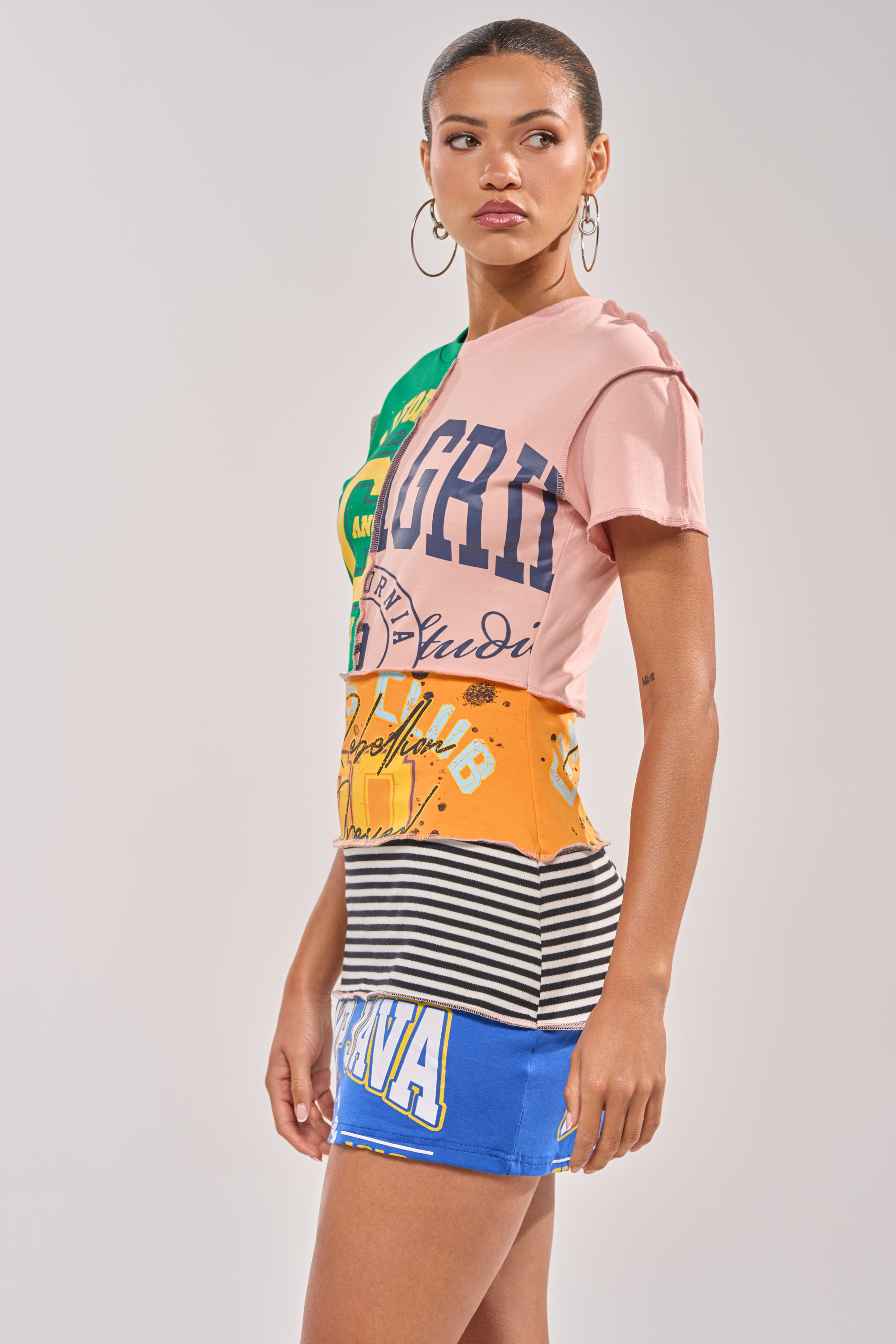 A woman stands sideways in the LEVEL UP GRAPHIC MINI DRESS, featuring graphic prints, stripes, and bold text. She wears hoop earrings, her hair is tied back, and she looks over her shoulder with a neutral expression.