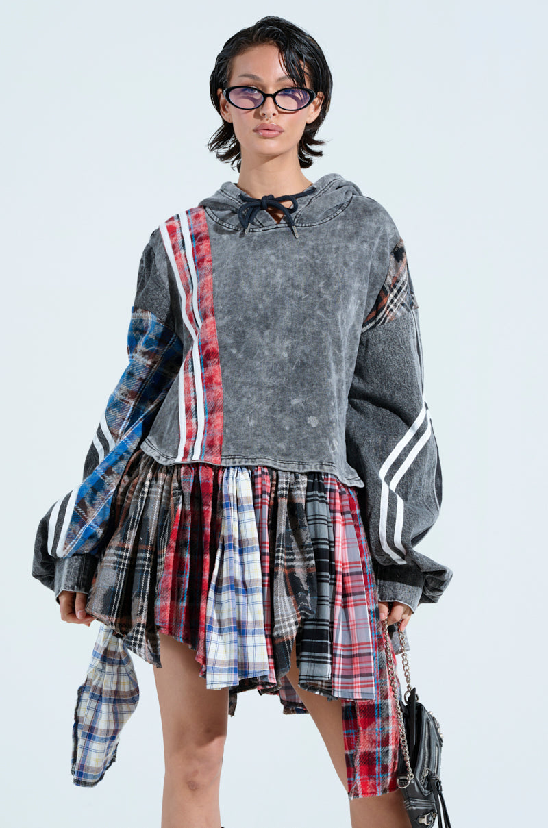 A person wearing glasses models the "A LITTLE BIT OF EVERYTHING PLAID SWEATSHIRT DRESS," featuring an oversized gray hoodie with white stripes. They hold a small handbag, blending cozy cotton with grunge styles for a casual yet edgy look.
