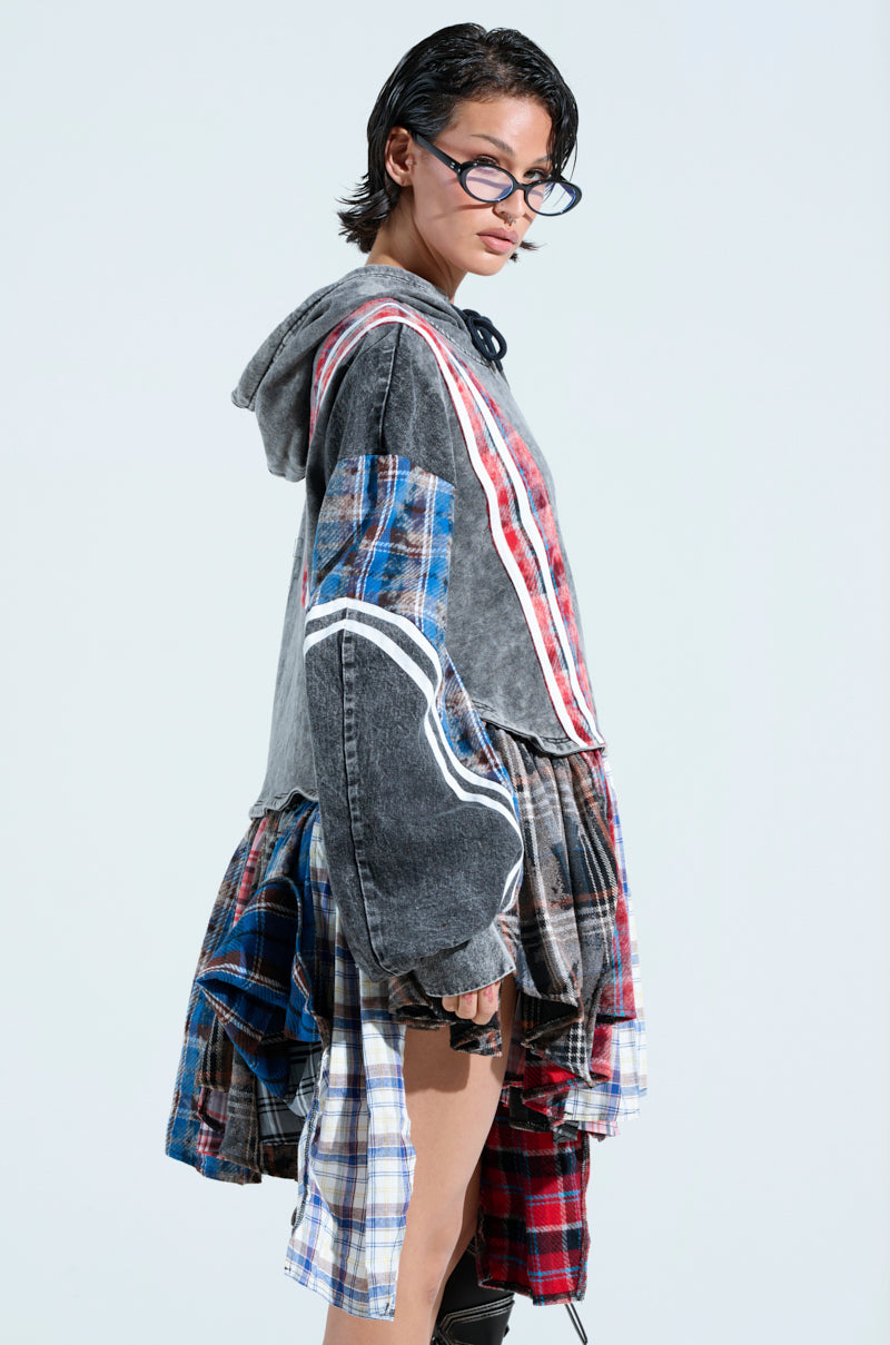 A person with short dark hair and glasses models the A LITTLE BIT OF EVERYTHING PLAID SWEATSHIRT DRESS, featuring a cozy cotton hoodie with an asymmetrical design and layered look. Patchwork plaid combines with denim in an oversized fit against a plain light background, highlighting its eclectic style.