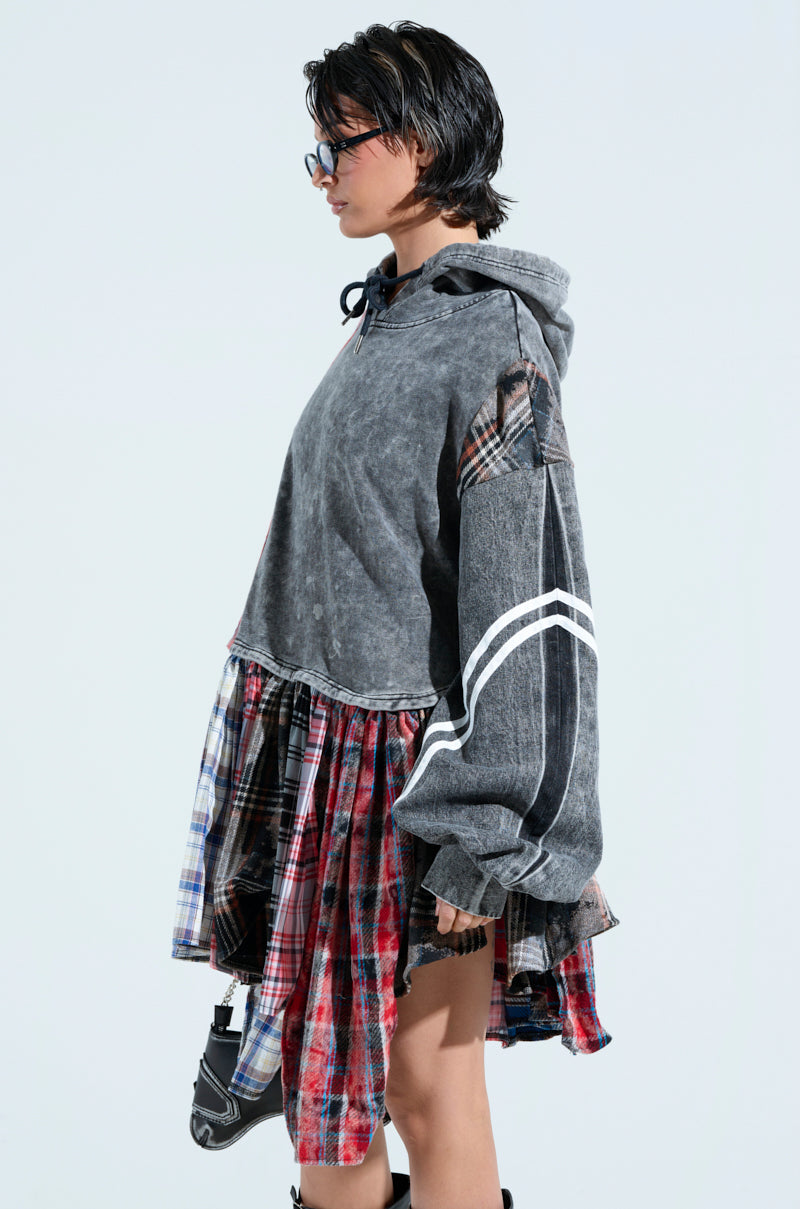 A person with short dark hair and glasses stands in profile wearing the A LITTLE BIT OF EVERYTHING PLAID SWEATSHIRT DRESS, featuring mismatched plaid sleeves and skirt, in an oversized cozy cotton gray design. They clutch a small purse against a plain light-colored background.