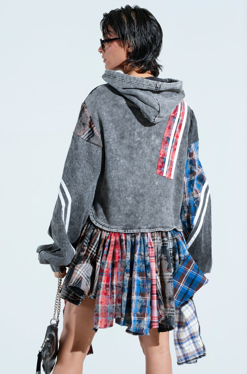 A person stands with their back to the camera in an A LITTLE BIT OF EVERYTHING PLAID SWEATSHIRT DRESS, featuring colorful striped patches and a plaid design. They hold a chain-strap bag and wear sunglasses, showcasing an oversized fit that blends cozy cotton textures and bold patterns seamlessly.