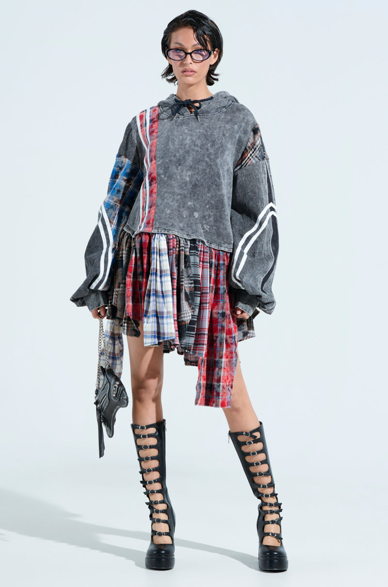 A person models the "A LITTLE BIT OF EVERYTHING PLAID SWEATSHIRT DRESS," featuring an oversized fit with gray, red, and blue plaid panels. It's styled with a plaid skirt and knee-high black boots with buckles on a plain white background.