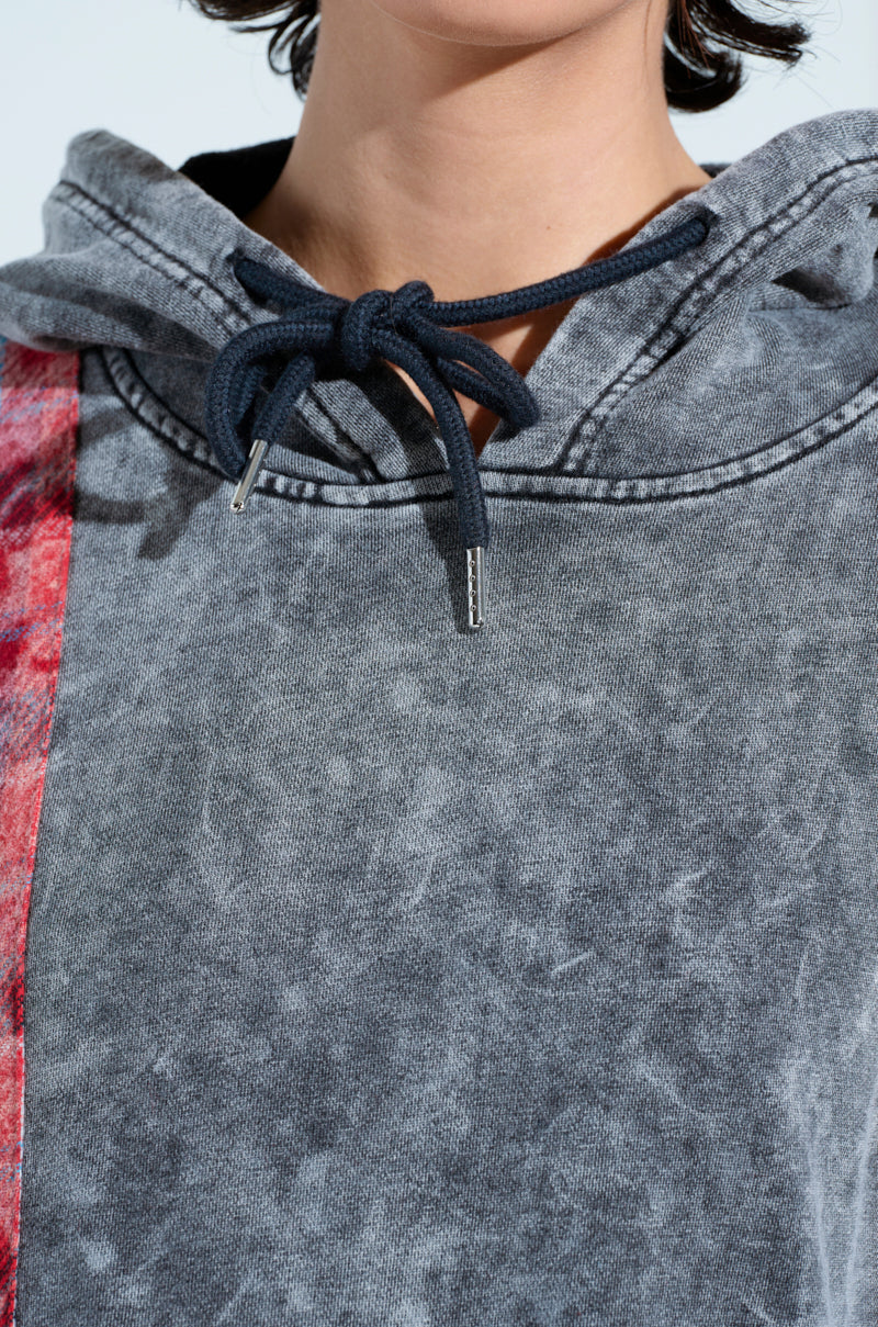 A person wears a gray, textured A LITTLE BIT OF EVERYTHING PLAID SWEATSHIRT DRESS with an oversized fit and drawstring. A red plaid stripe adorns the left side. The focus is on the upper part of the dress, with the person's head partly visible.