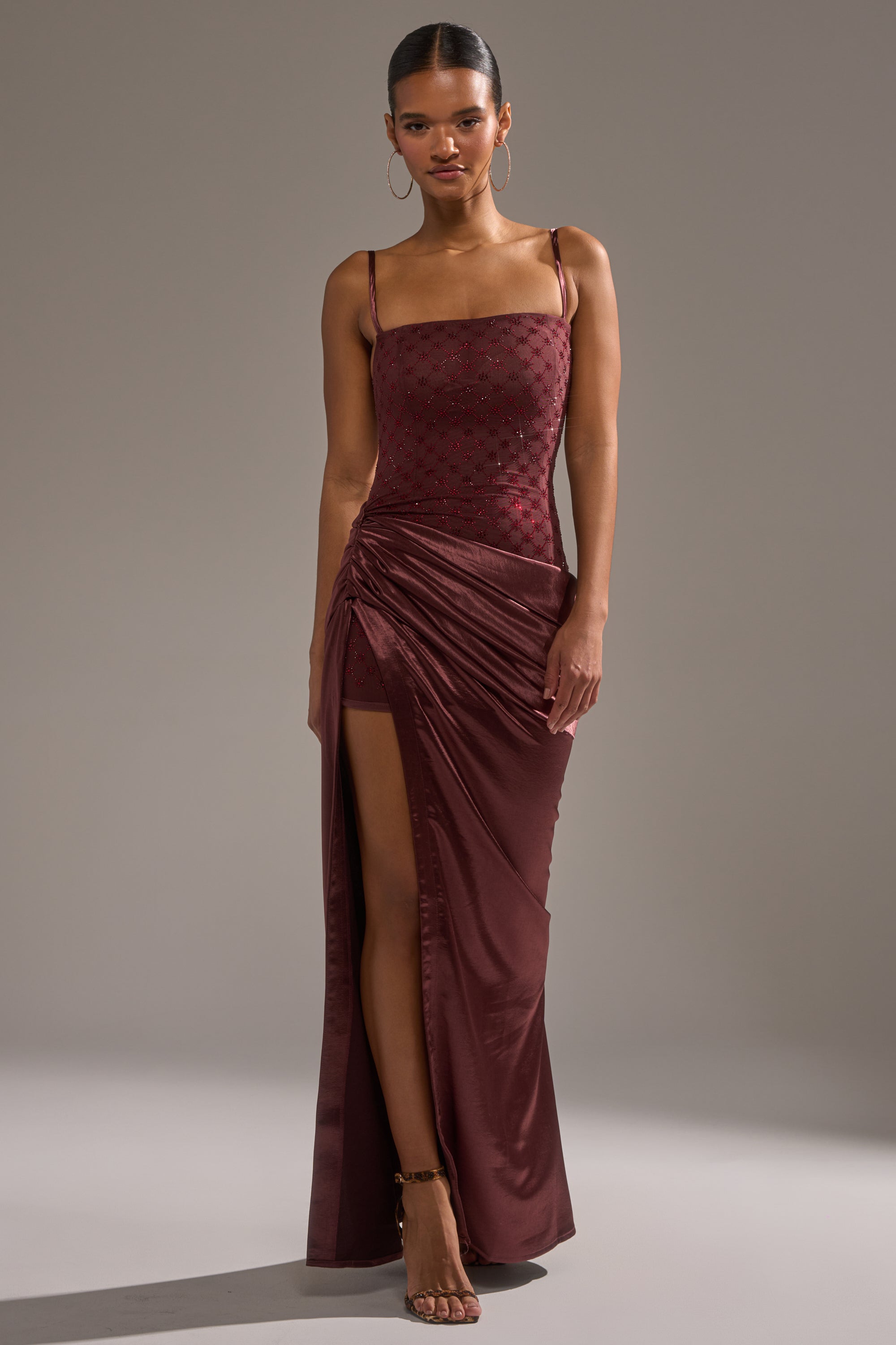 Wearing the RUNAWAY SATIN MESH MAXI DRESS featuring a patterned bodice, fitted silhouette, high side slit, and ruched details, a woman stands confidently with hoop earrings and ankle-strap heels against a plain background.