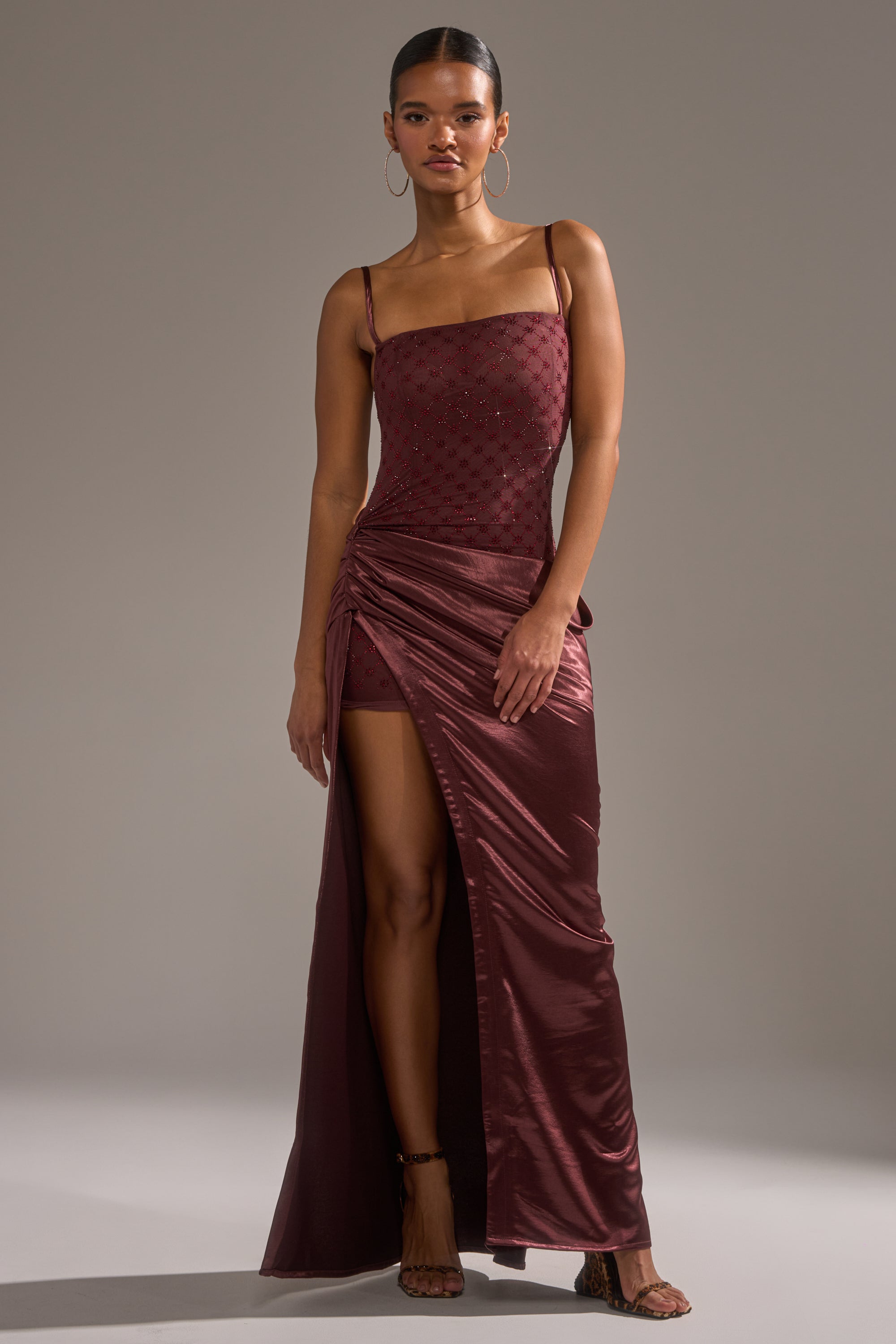 A woman stands confidently in heels against a plain gray background, wearing the RUNAWAY SATIN MESH MAXI DRESS—sleeveless with a square neckline, beaded bodice, high slit, ruched detail, and burgundy satin mesh fabric.