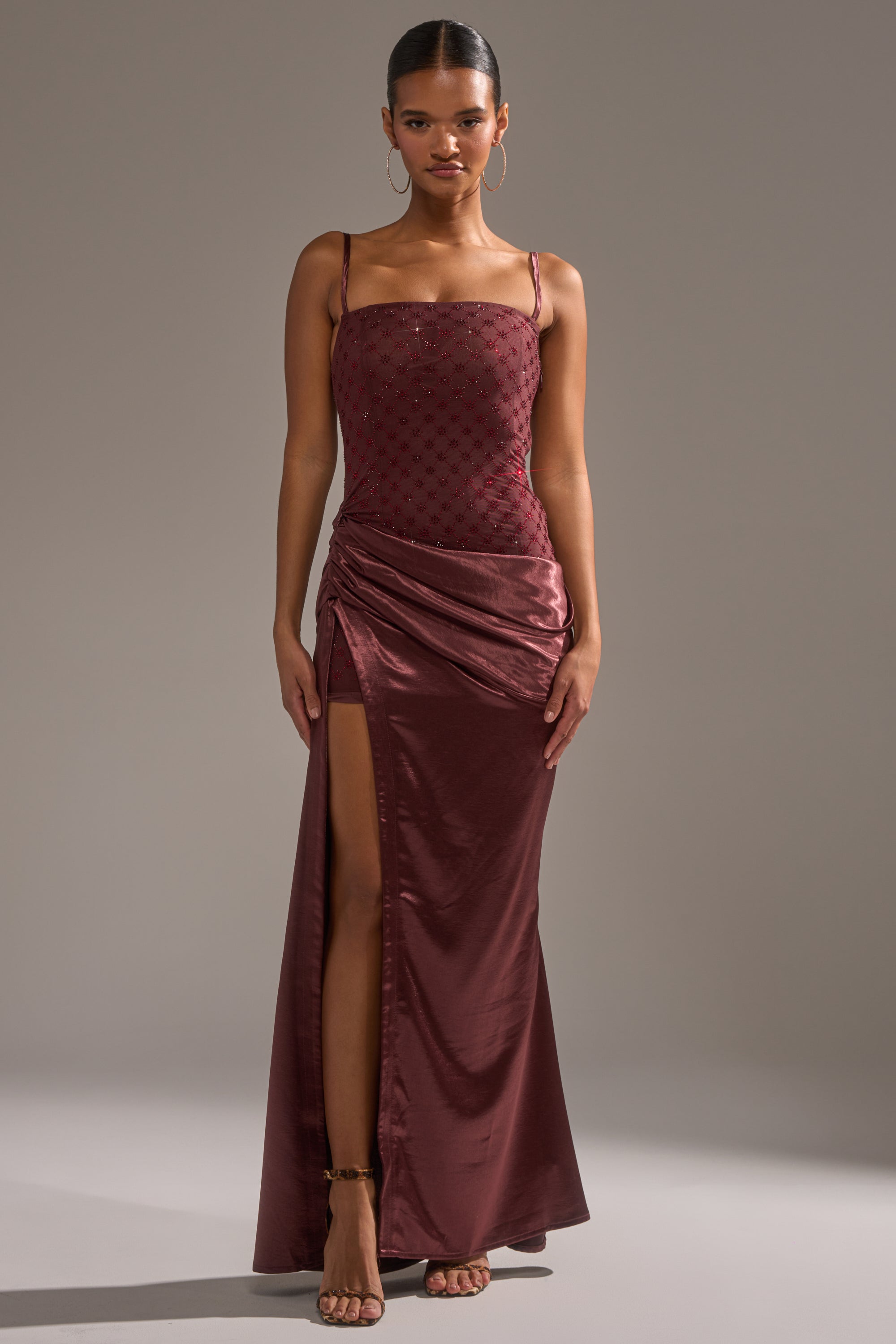A woman stands facing forward against a neutral background, wearing the RUNAWAY SATIN MESH MAXI DRESS—a sleeveless burgundy maxi with thin straps, ruched detailing, mesh accents, and a side slit.