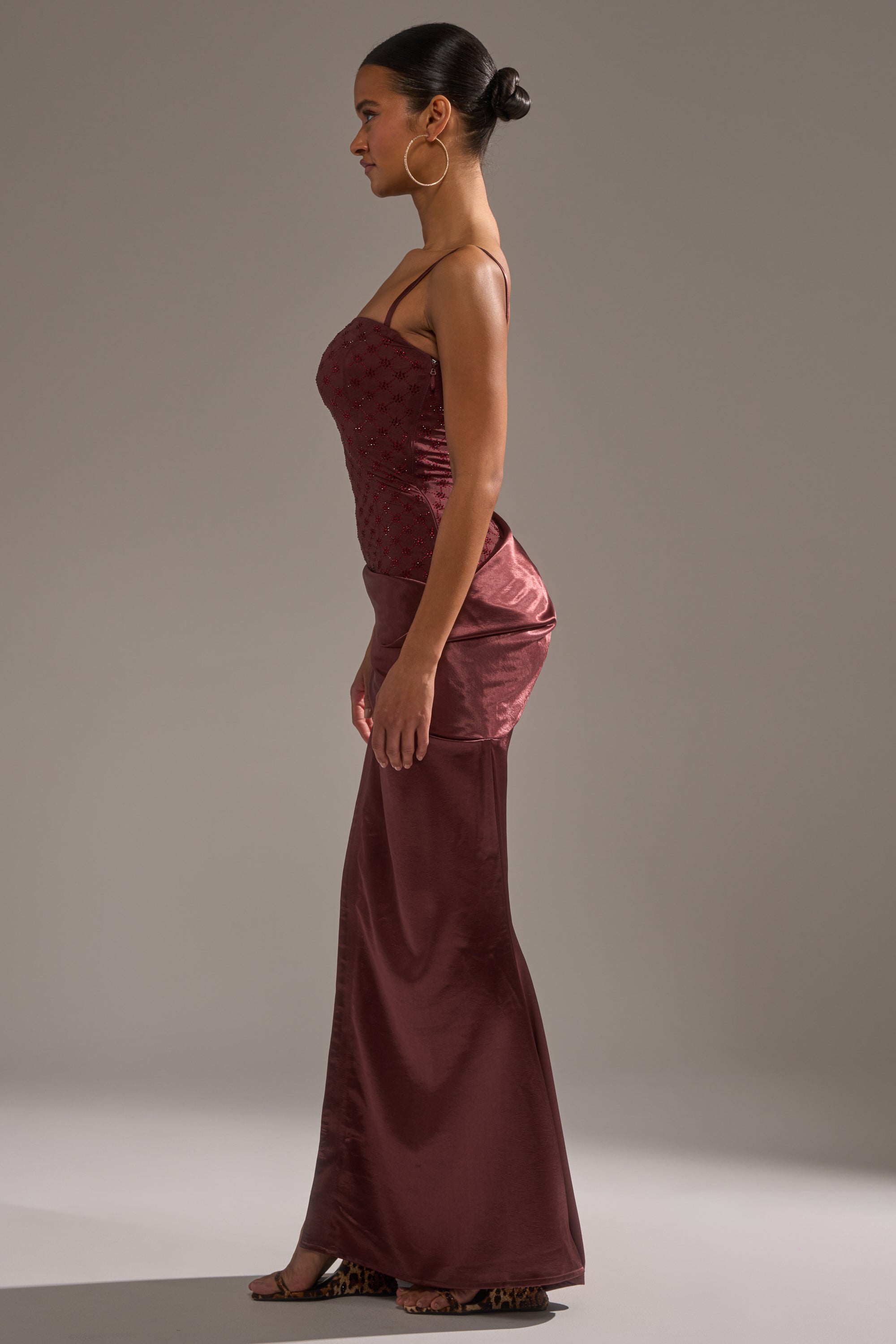 A woman stands in profile against a plain background, wearing the RUNAWAY SATIN MESH MAXI DRESS in burgundy. The sleeveless, form-fitting maxi features a satin mesh finish. She completes the look with a neat bun, large hoop earrings, and heels.