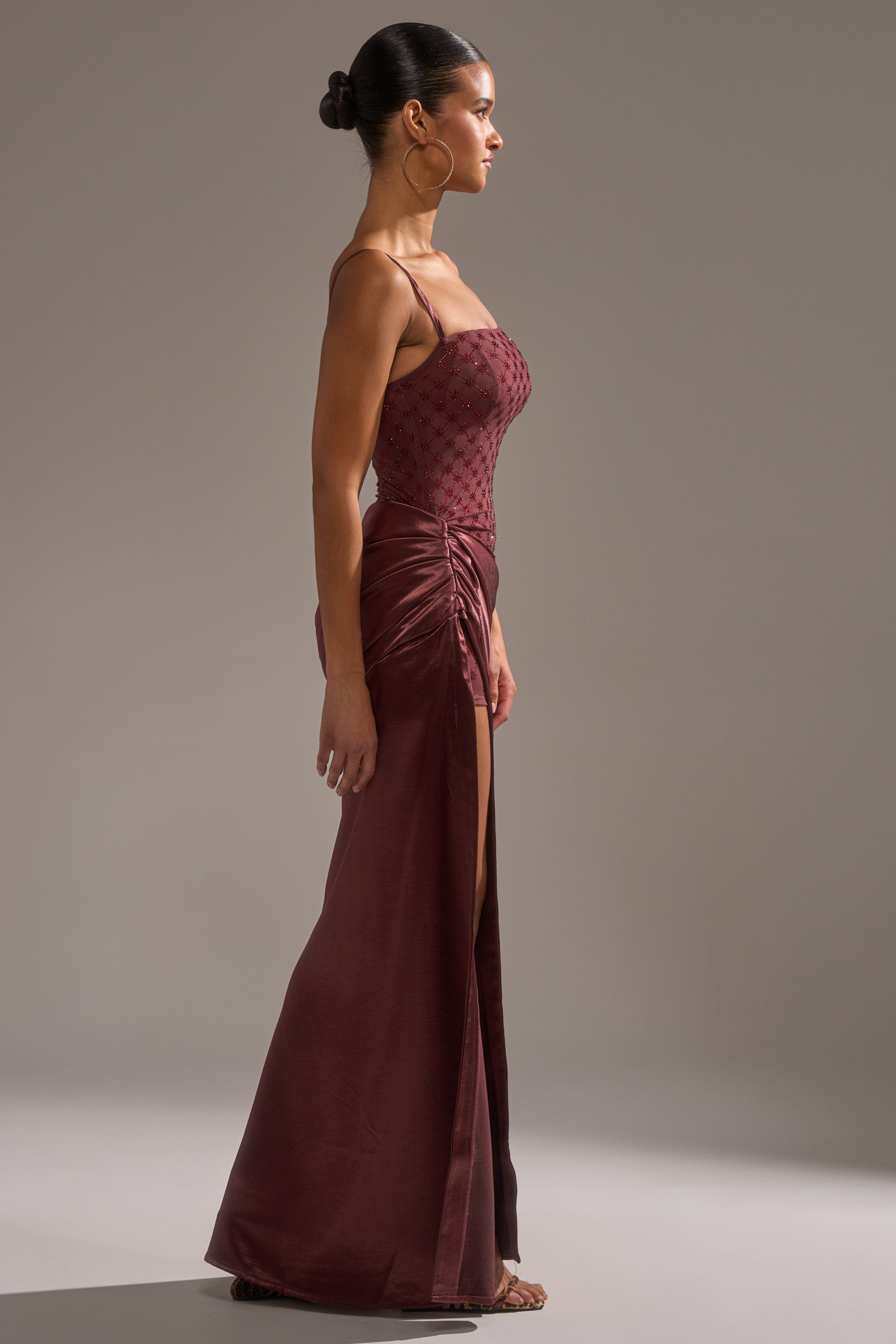 A woman in profile wears the RUNAWAY SATIN MESH MAXI DRESS—sleeveless, burgundy, with a patterned fitted bodice, thin straps, ruched hip detail, and high slit. Her hair is styled in a neat bun against a plain background.